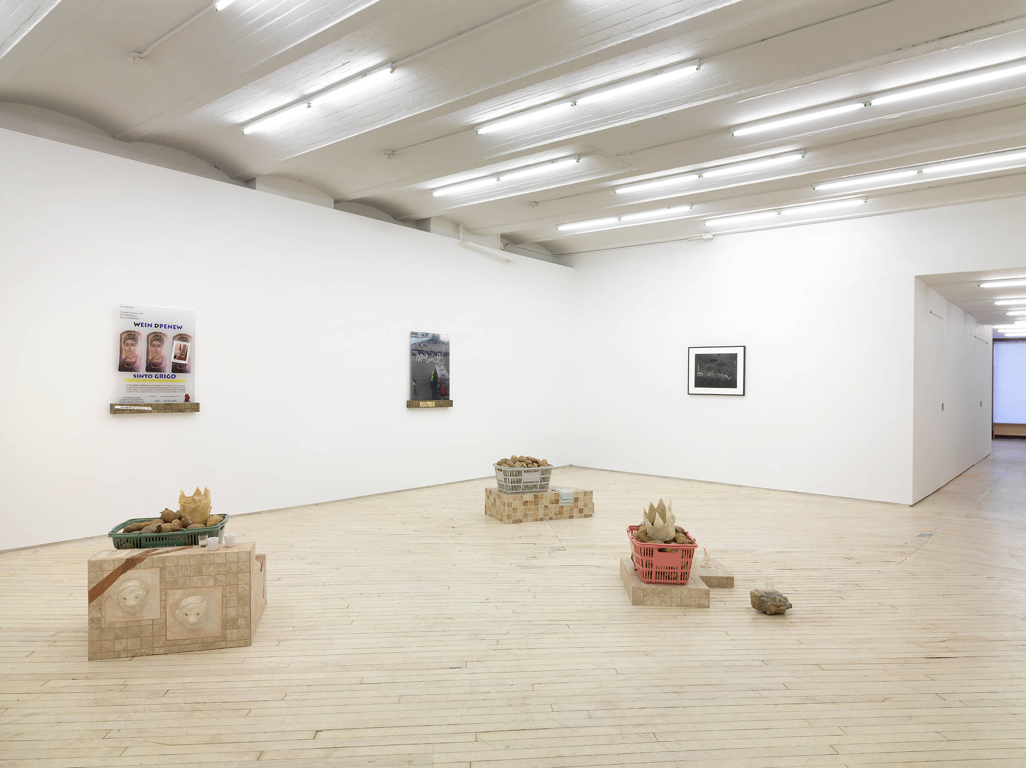 Installation view of three sculptures by Libby Rothfeld are on the floor, all are made using ceramic tiled plinths and plastic shopping baskets filled with real potatoes. Two pieces hang on the left wall that are also by Libby Rothfeld showing prints of hand made wine labels on a tiled shelf. One black and white photograph by Erica Baum depicting chalkboards hangs on the back wall of the gallery.