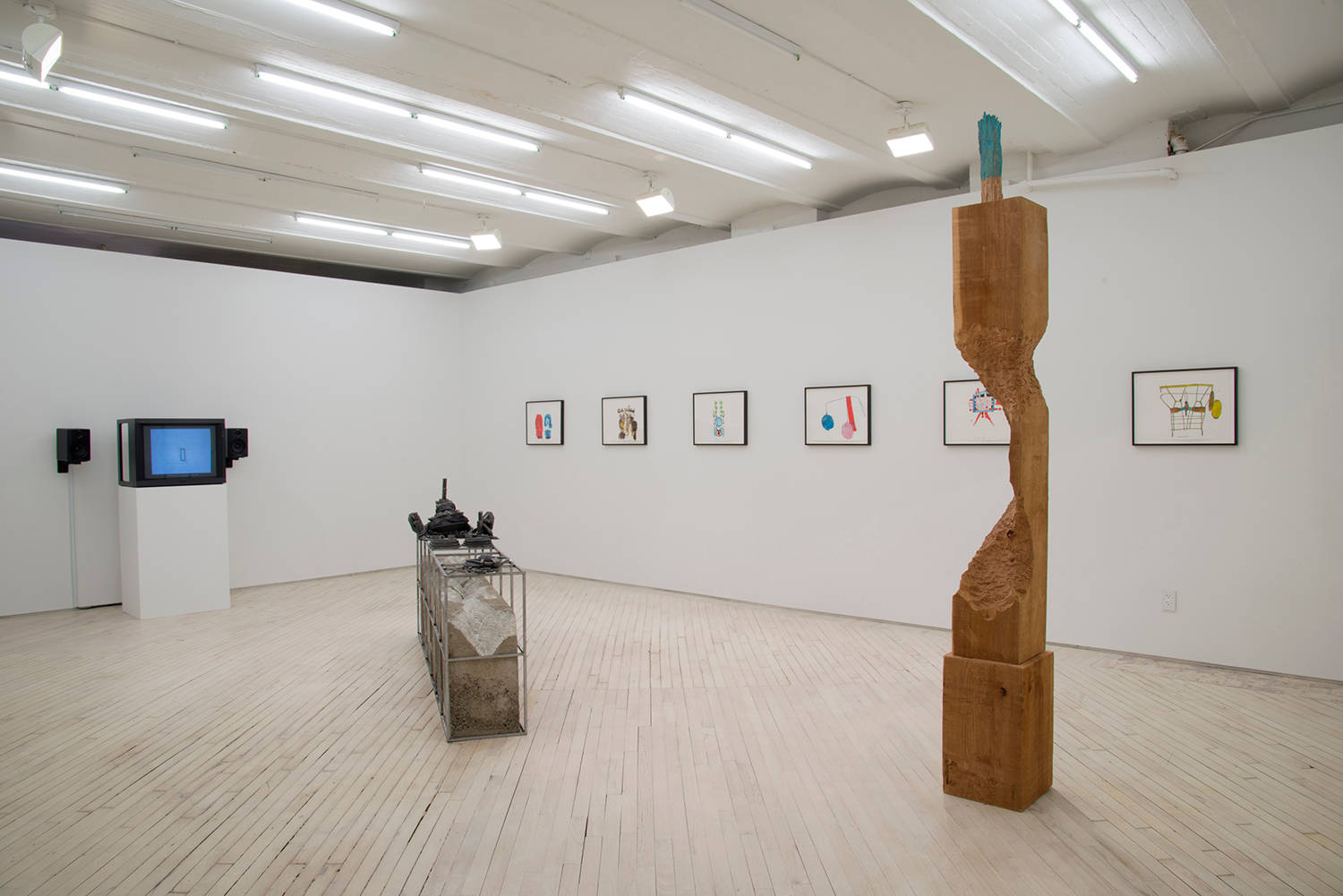 Installatoin view of the exhibition in the main space. At left, a monitor on a plinth plays a video by Wojciech Bakowski, at center a low concrete and metal sculpture by Lionel Maunz, on the right wall a suite of colorful ink drawings by Christine Rebet and in the foreground a roughly carved tall wooden sculpture by David Adamo.