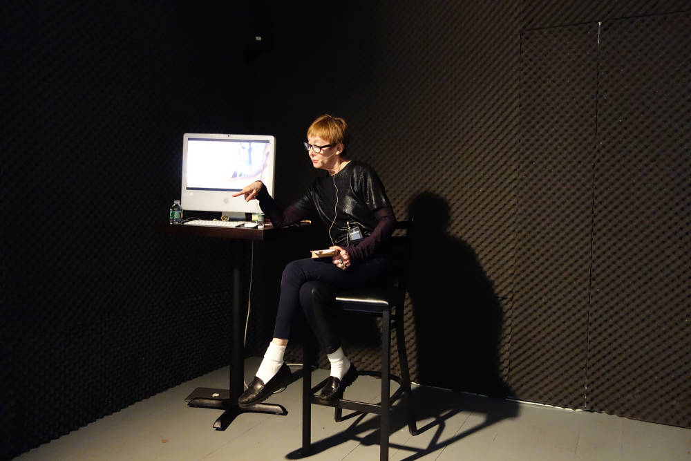 Image of a sound-proofed room walled with egg crate foam: a red headed woman wearing black sits on a stool next to a computer on a tall table. She is wearing a small microphone and is pointing her right finger out of the frame.