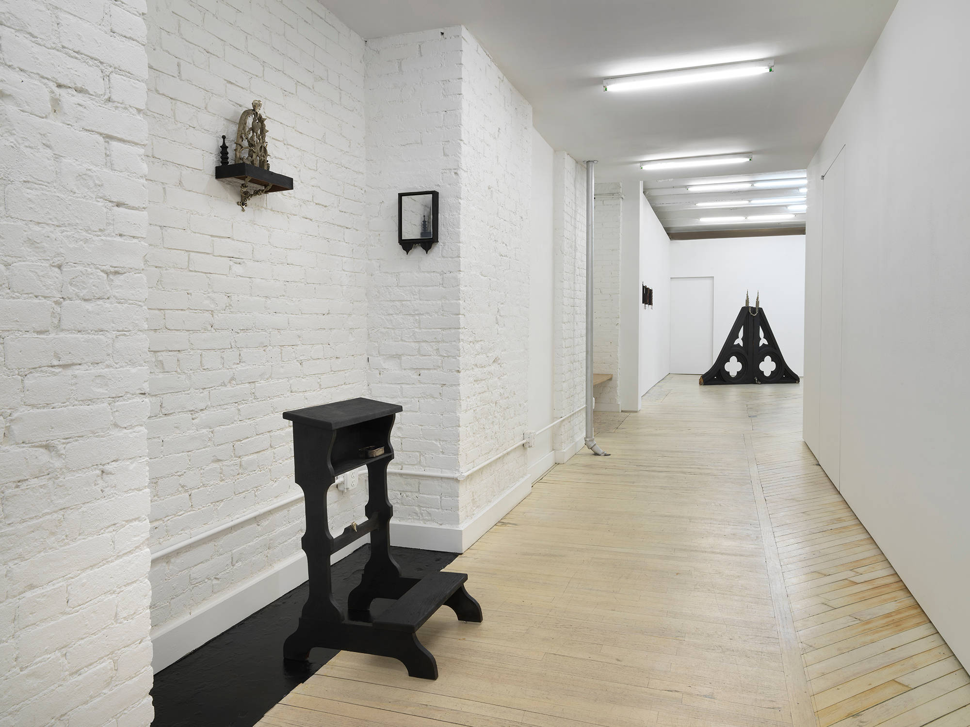 Installation view looking down the hallway, a black sculptural installation comprising a black chapel kneeler and a two small wall works hang on the brick wall at left. At the end of the hallway a large black pyramid shape sculpture stands at the end of the hall in the main space.
