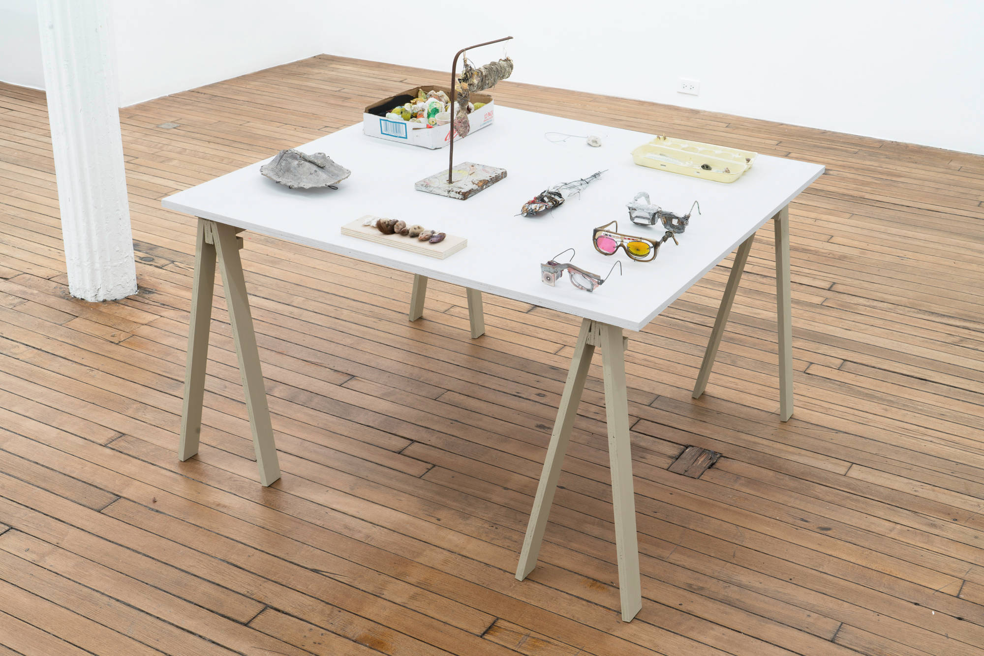 A white tabletop sits on beige sawhorse legs. The tabletop contains sculptures dating back 20 years by the artists Matt Hoyt and Tom Thayer.