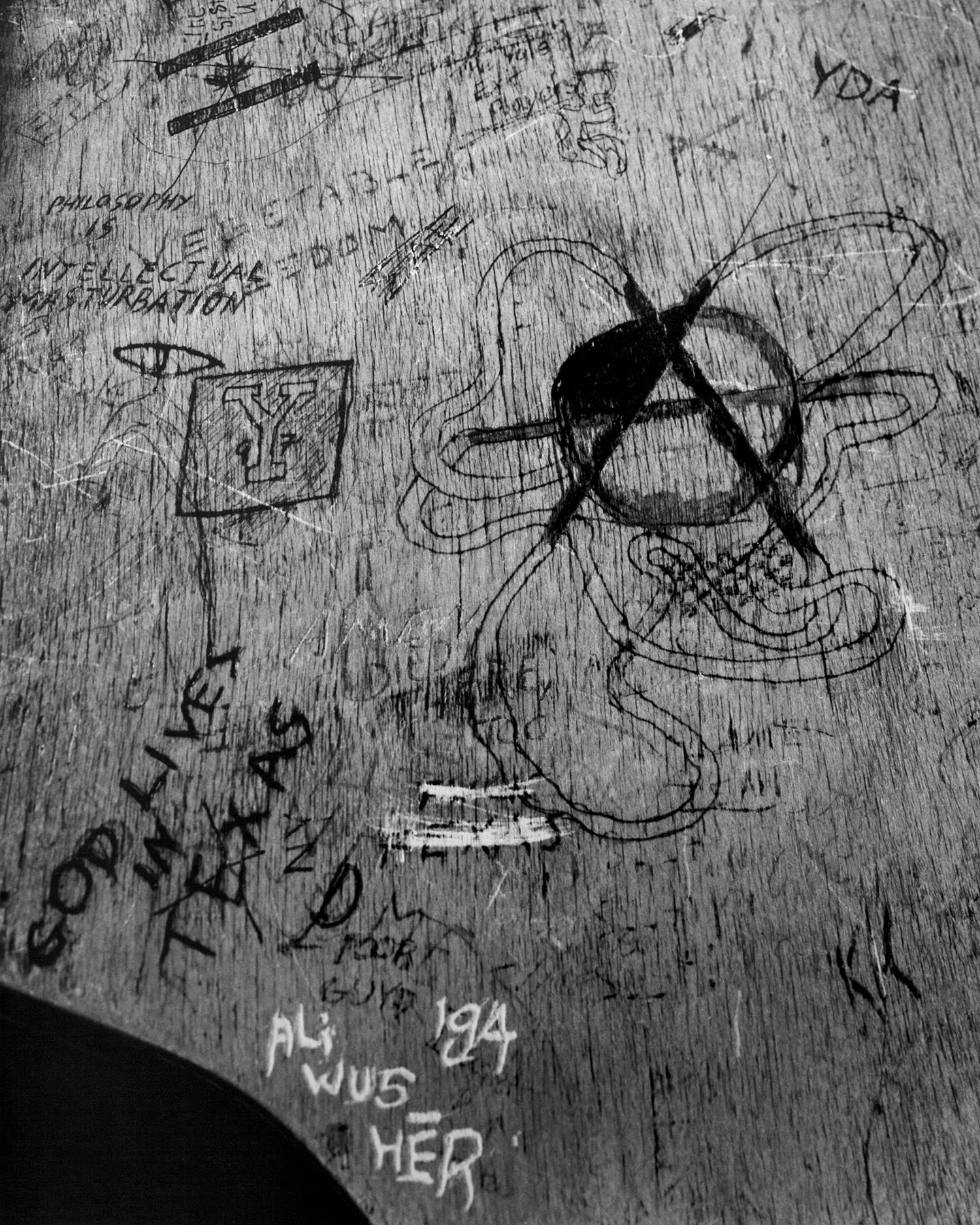 Black and white photograph of a section of a carved wooden school desk top. Various words are carved and drawn into it. A large anarchy symbol is visible on the right hand side with a snaking knot pattern coming out of the lines of the letter A. We see a canadian flag to the top left and the words 'God Lives in Texas' and 'Ali wus hēr '84' on the bottom left.