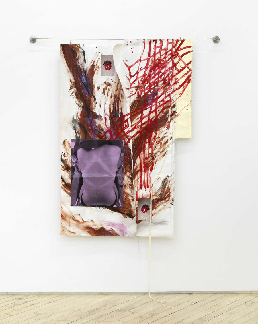 An abstract painting of irregular shape hung from metal hardware. The painting has multiple layers of cutout fabric. The dominant colors are red, purple and brown. 