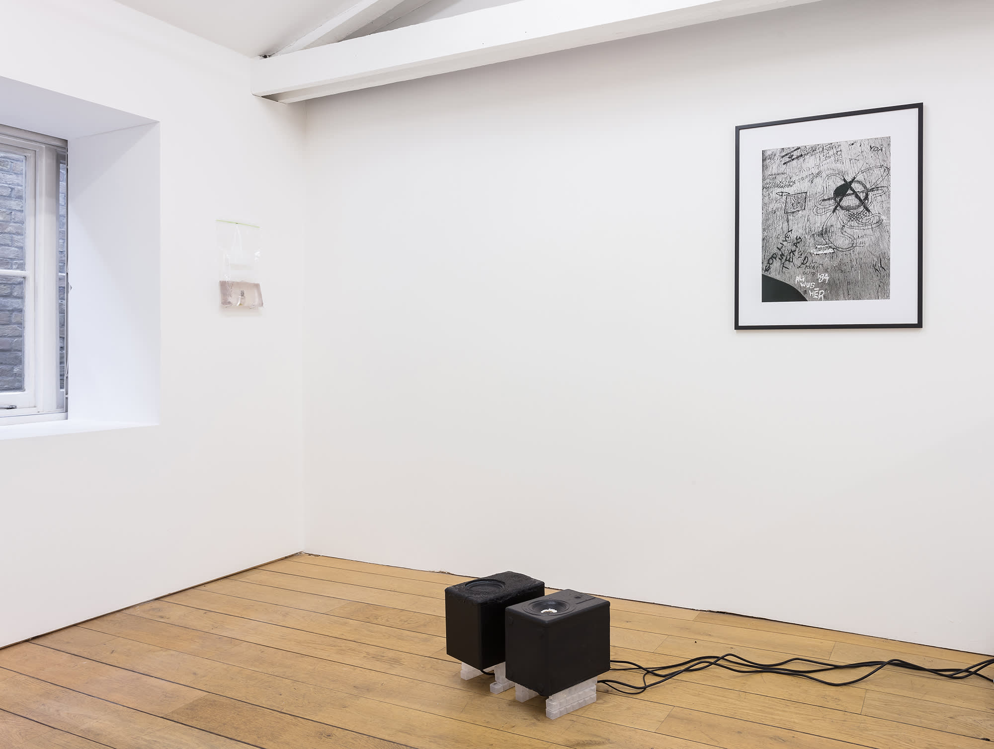 Installation view of a gallery with wooden floors and a slanted ceiling, partial view of a window on the left. On the left wall next to the window is a plastic bag with liquid pinned to the wall. On the right wall is a black and white photograph in a black frame and white matte, of a carved school desk - an anarchy symbol is visible. On the floor are two black speakers with pearls inside the cones of the speakers and black cables trailing off to the right.