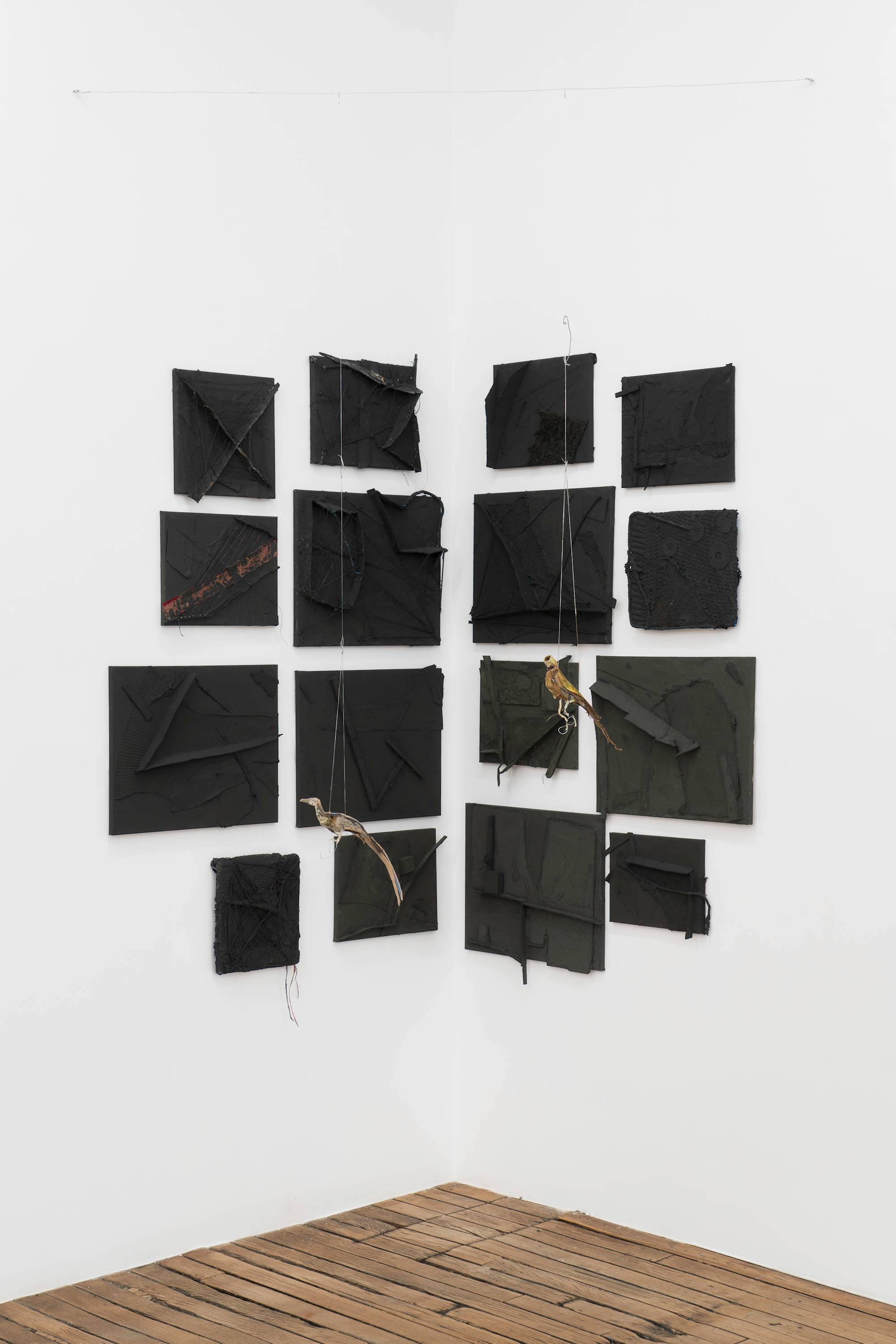 A group of small black paintings, textured with cardboard and other material, hung in the corner of a gallery above a wood floor. 2 bird sculptures are suspended on string in front of the group of paintings.