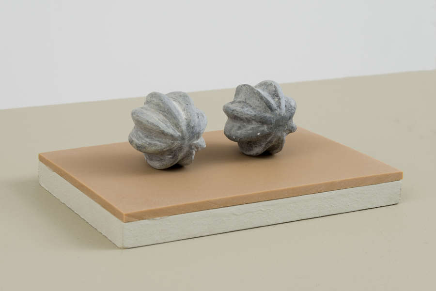 Two handmade sculptures resembling chestnuts resting on top of an orange-tinted plinth with a white wall behind them partially in view. 