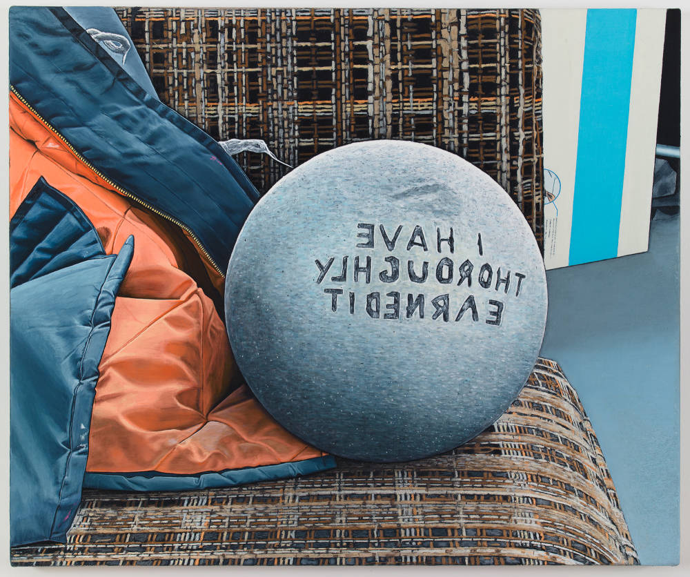Oil on canvas painting depicting a gray sphere sitting on a plaid couch with the words "I HAVE THOROUGHLY EARNED IT" carved in reverse on the sphere, which is also resting partially on a black and orange flight jacket. 