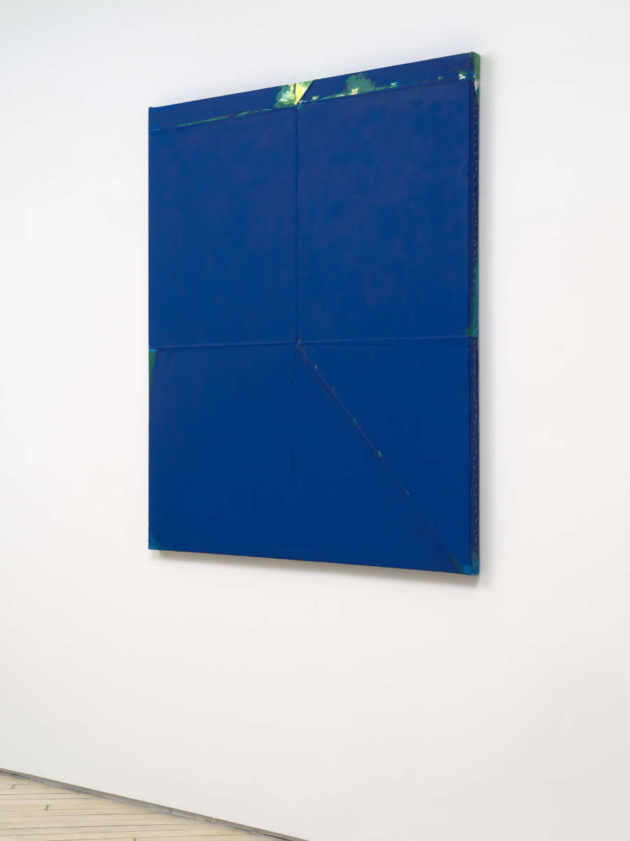 A sideview of a large blue monochromatic painting with a green accent at the top. 