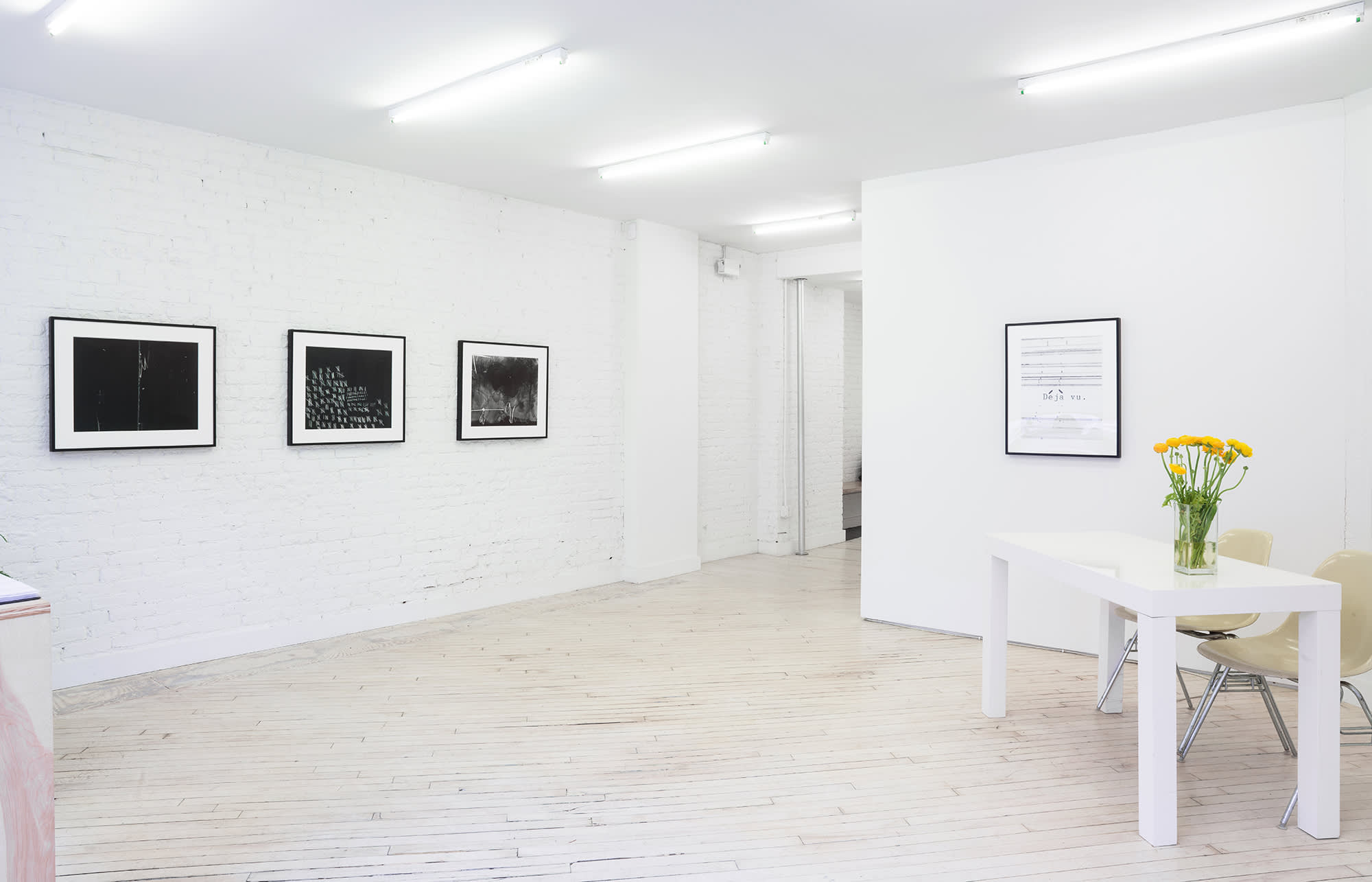 Image of the white front room of the gallery. At left 3 framed black and white photos, predominantly all black with some white marks, hang on the brick wall. At left a single framed black and white photo hangs, mostly white with black lines and the black typewritten 'Déjà vu.' There is a white desk at the right, and yellow flowers in a vase.