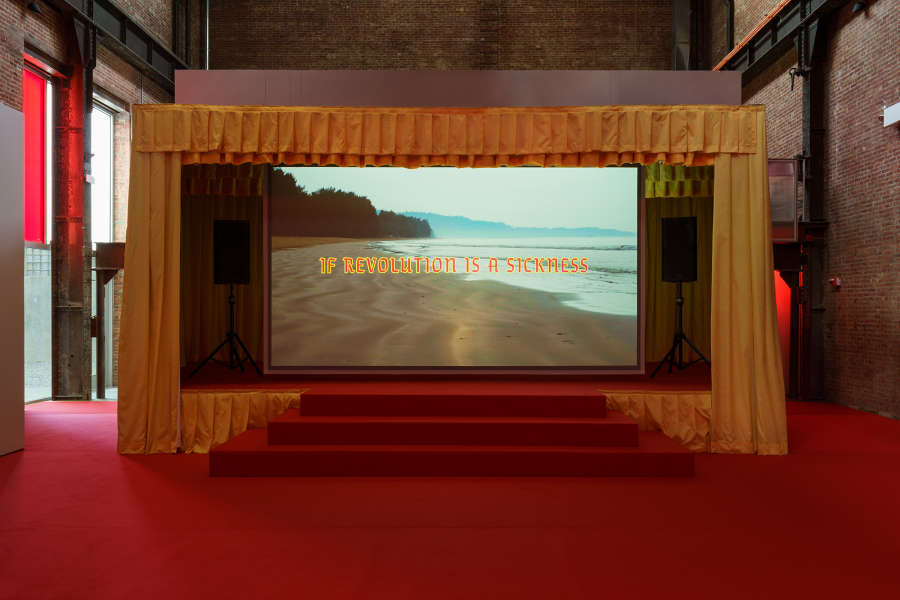 A darkened brick gallery space with an installation resembling a red carpeted concert stage with yellow curtains. In the center of the stage is a large video projection of a coastline with the words "IF REVOLUTION IS A SICKNESS."