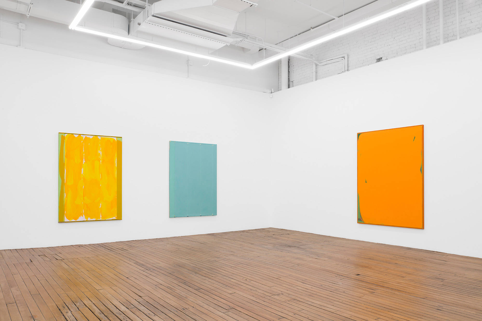 Three larges monchromatic paintings hung in the corner of a large gallery rendered primarily in yellow, blue, and orange.