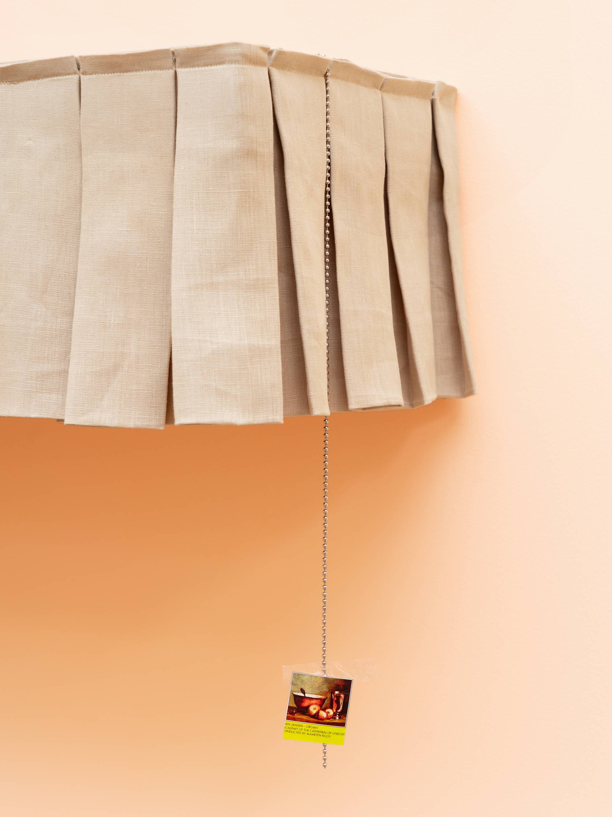 Detail of a sculptural shelf hanging on a peach colored wall. The shelf is lined with a short, beige, pleated curtain that is parted to the left of center by two vertical, yellow cushions, wrapped in cellophane, and an orange shoe box. A small picture hangs from the right side of the shelf by a ball chain.