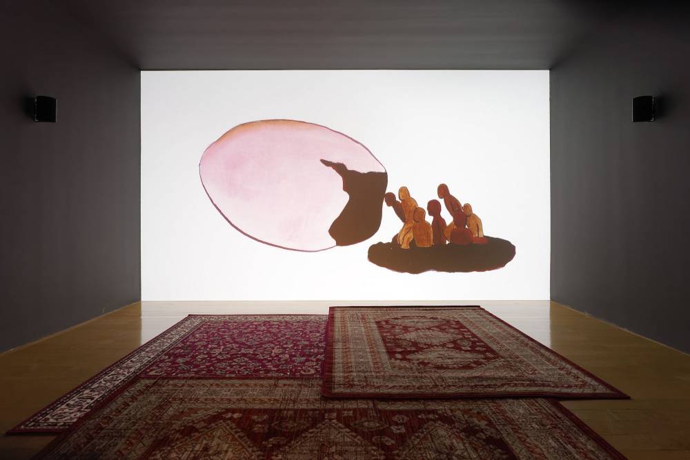 A large projection of an animation depicting several human figures entering a suspended egg shell. On the floor are large carpets. 