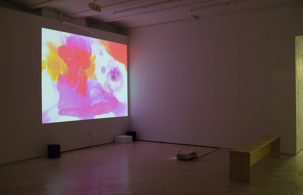 In a darkened gallery space, a video projection directly onto the wall containing orange, red, purple and pink blotches of color overlapping each other. A bench is sitting in front of the projection with the projector on the floor. 