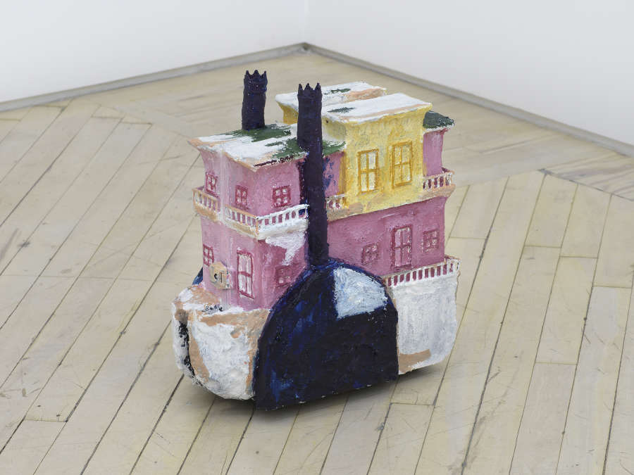 On the floor of a gallery space, a sculpture resembling a boat rendered in pink, yellow, blue, and white. 