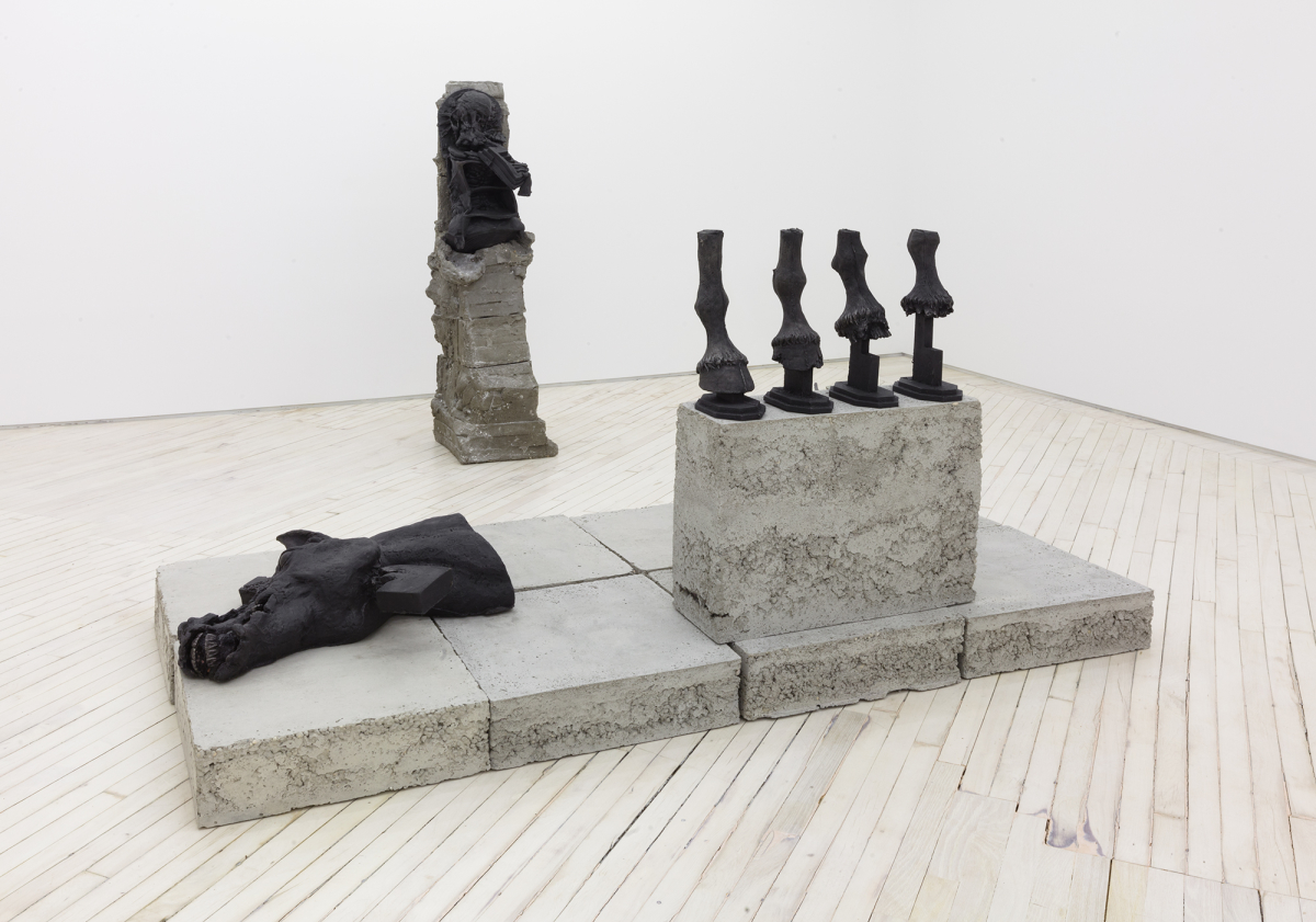 Installation view of cast iron and concrete sculpture resting on the floor, depicting a severed horse head and four horse hoofs standing.