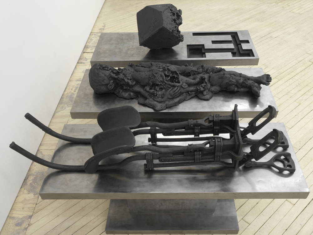In a gallery space, a large cast iron metal sculpture rests on the floor. The sculpture is made up of multiple components. Cast iron forms resemble a contraption device, an abstracted body, and a cubic form. 