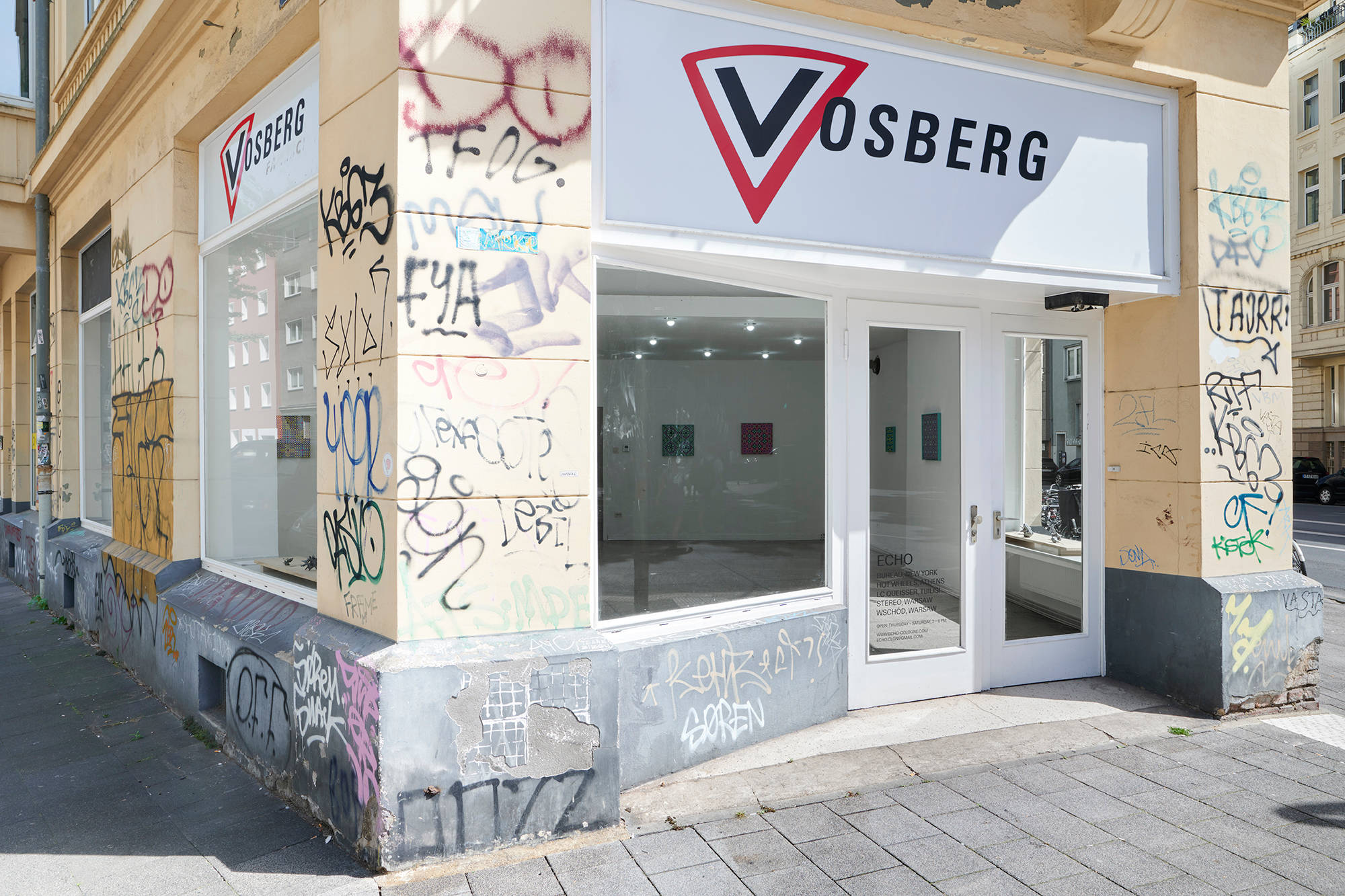 Outside view of the gallery with the word "Vosberg" on the top of the building, with a yellow wall that has graffiti covering it. Through two large windows, you can see the exhibition from outside. The door has the title of the gallery "Echo" and Bureau, New York.
