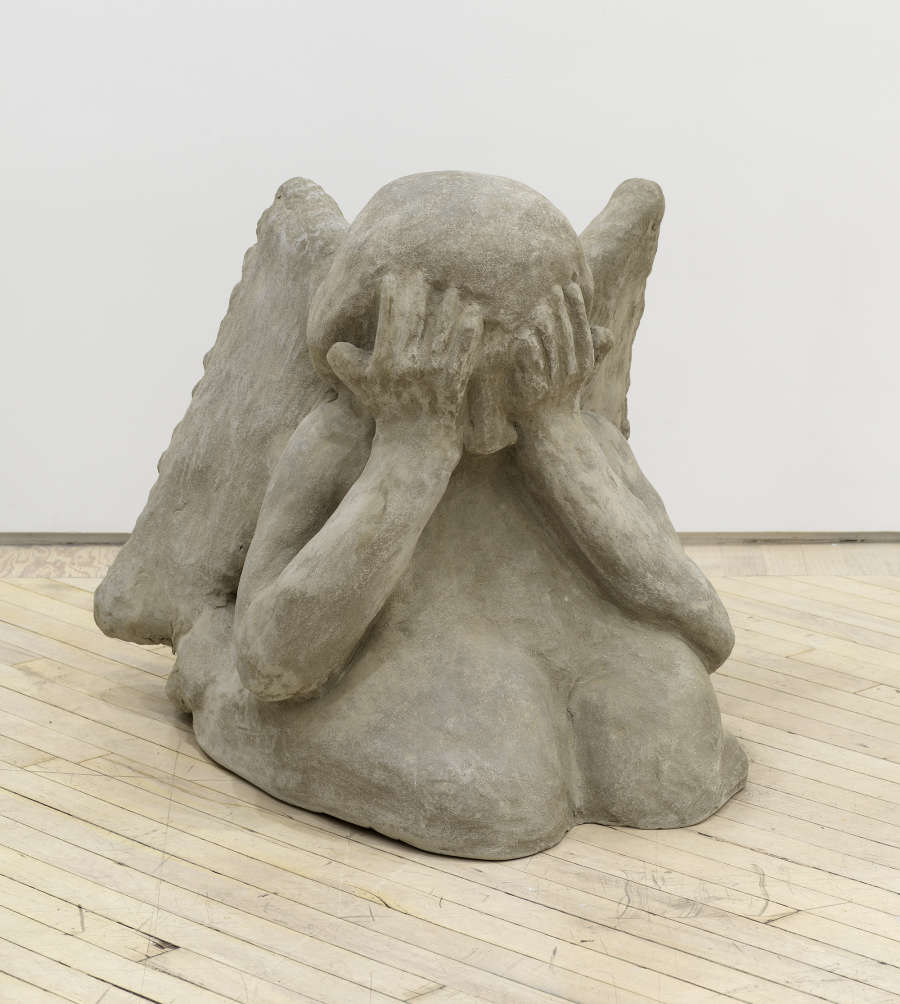 Image of an artwork by Libby Rothfeld of a cartoonishly rendered angel seated on the floor with wings and its hands covering its eyes. The angel is sculpted from concrete.