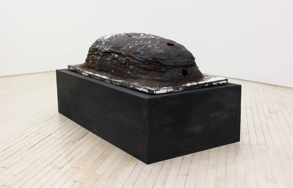 In the center of a gallery space, a large object resemble a tomb stone or grave. The sculpture has a rounded, dome-like structure on the top of the base. The object resembles gritty, industrial material. 