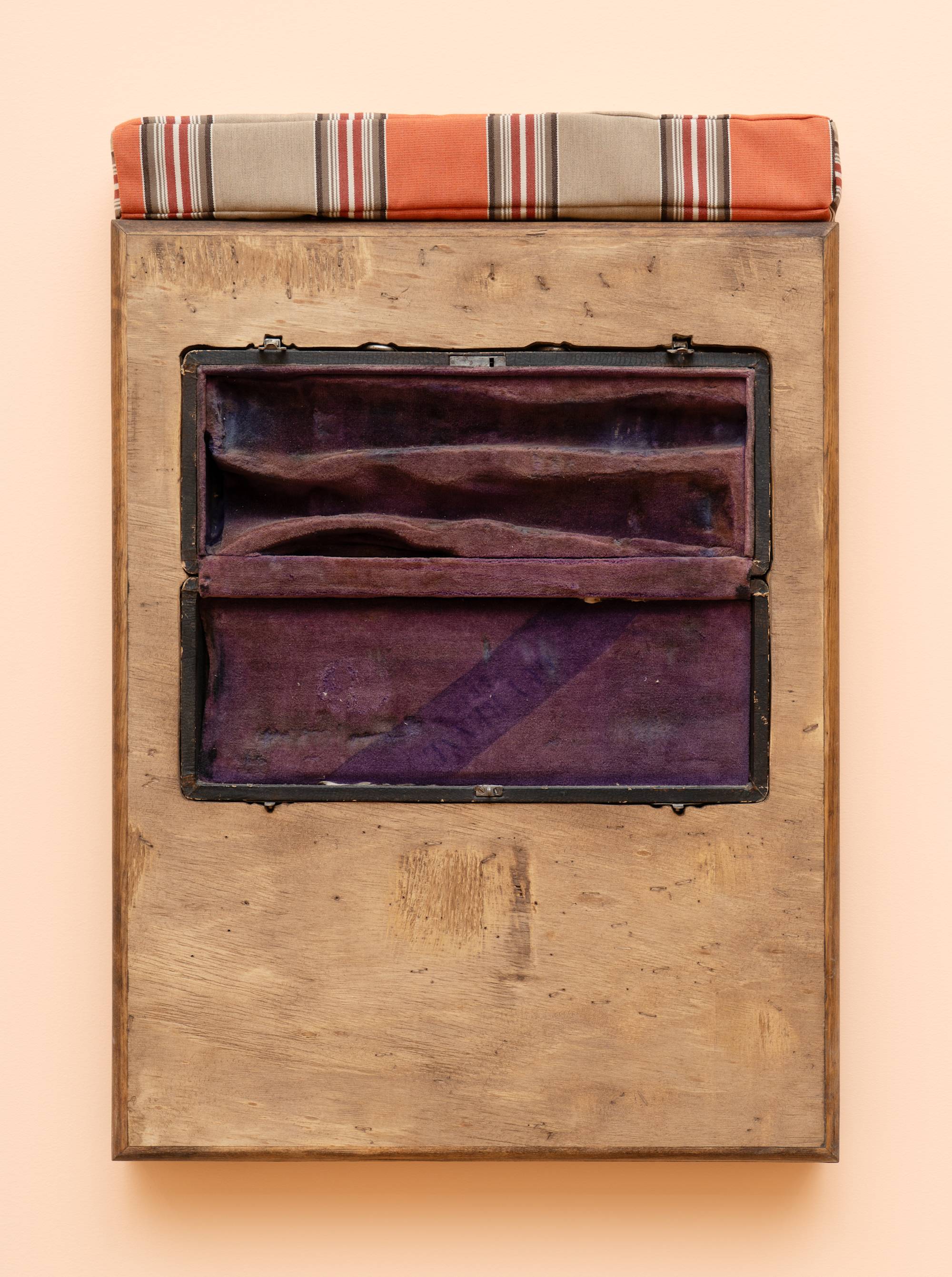 A wooden box sculpture with inset clarinet case with purple velvet lining, topped with a orange and beige striped cushion, hangs on a peach colored wall.