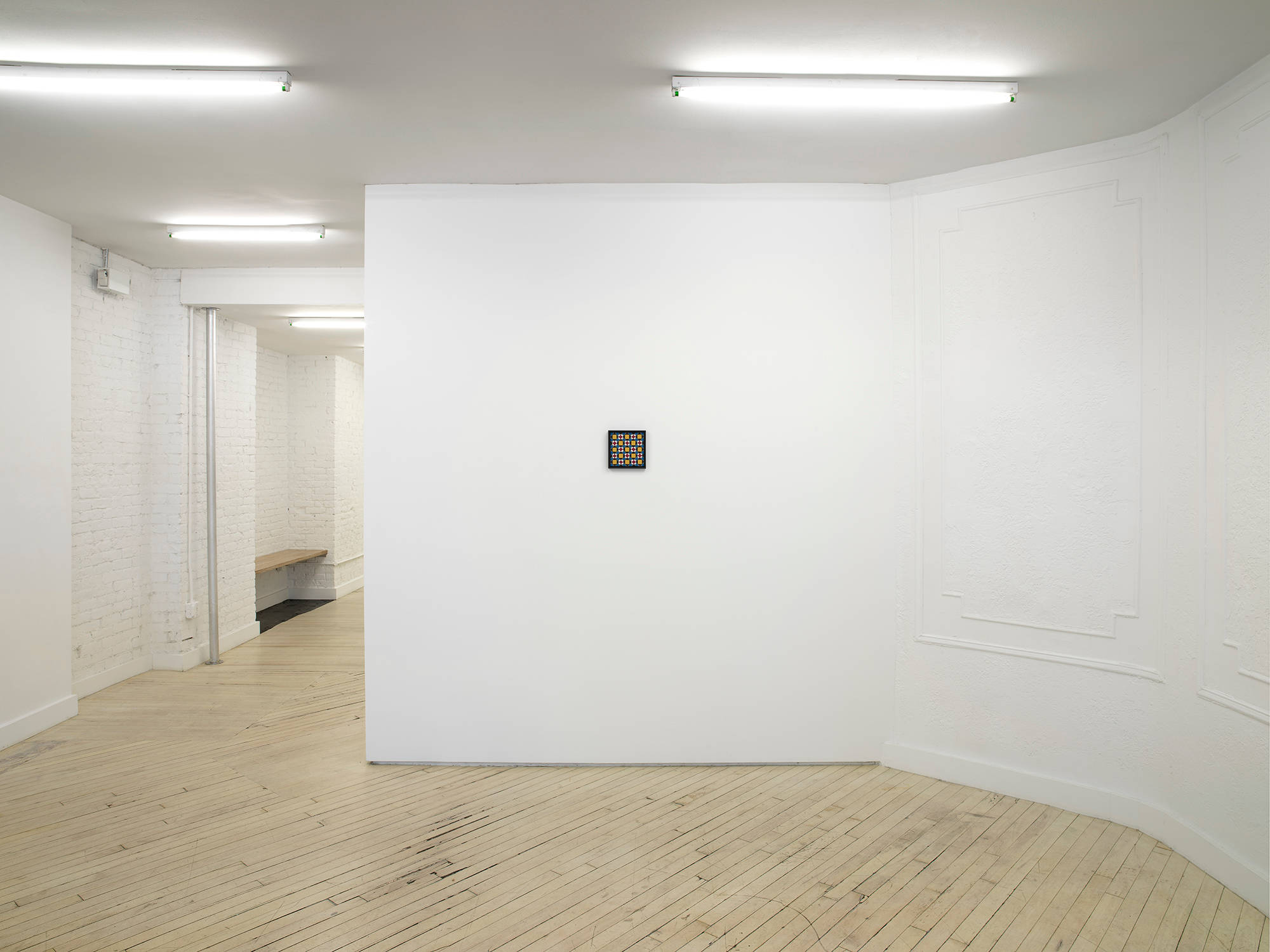 In the center of a gallery space, a small painting hangs in the middle of a white wall. The has a black frame and depicts a repeating grid pattern in blues, reds, and yellows. There is a hallway to the the left with a bench.