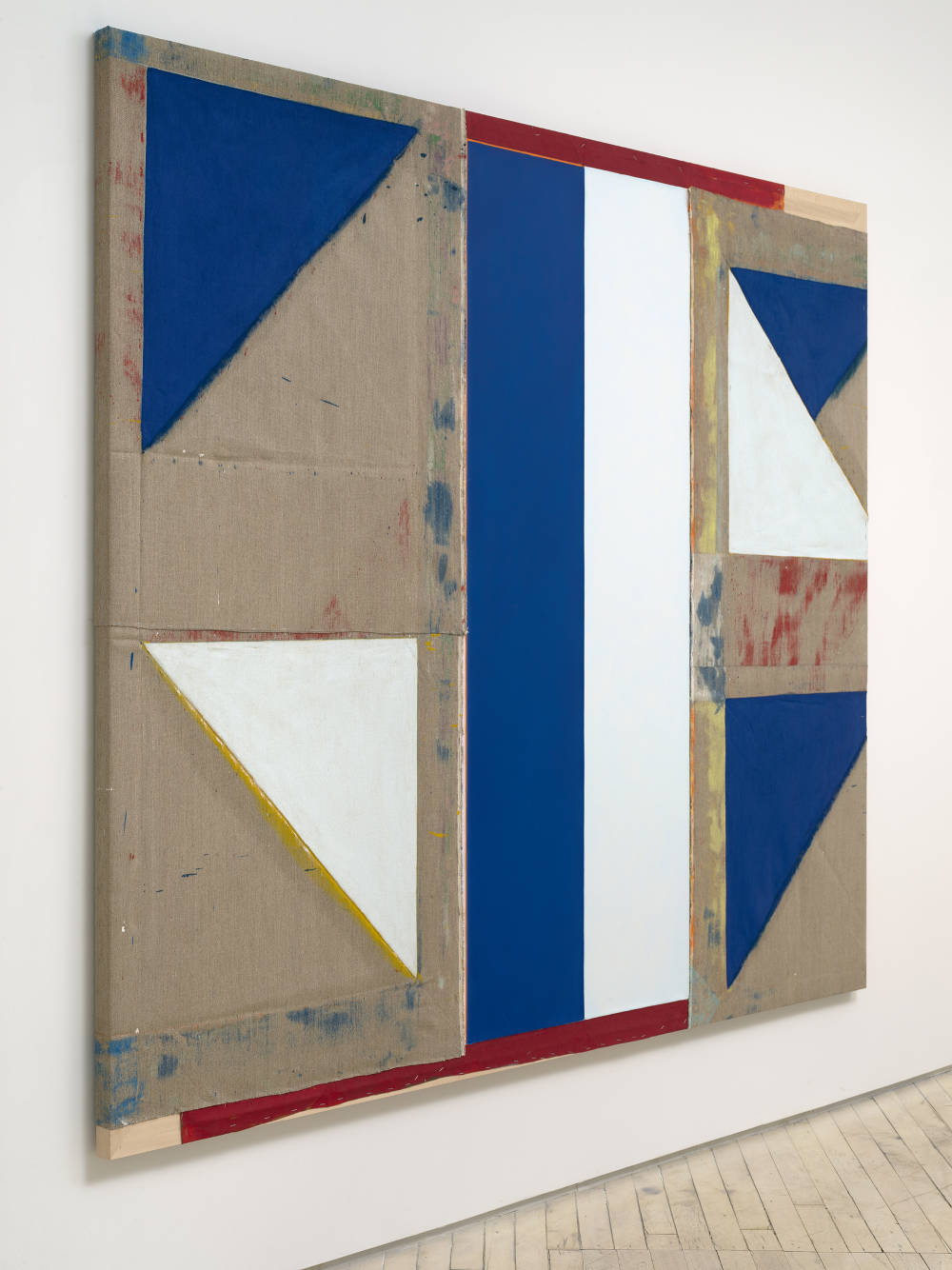 A side view of a large abstract painting. The painting consists of differently shaped areas of solid colors ranging from blue, red, white to brown. 