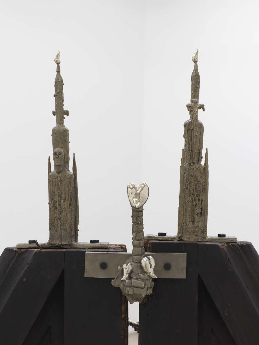 Detail of a sculpture's cast bronze elements in the form of pointy steeples and a brick chimney with a polished heart at the top, all affixed to a larger charred wooden structure below.