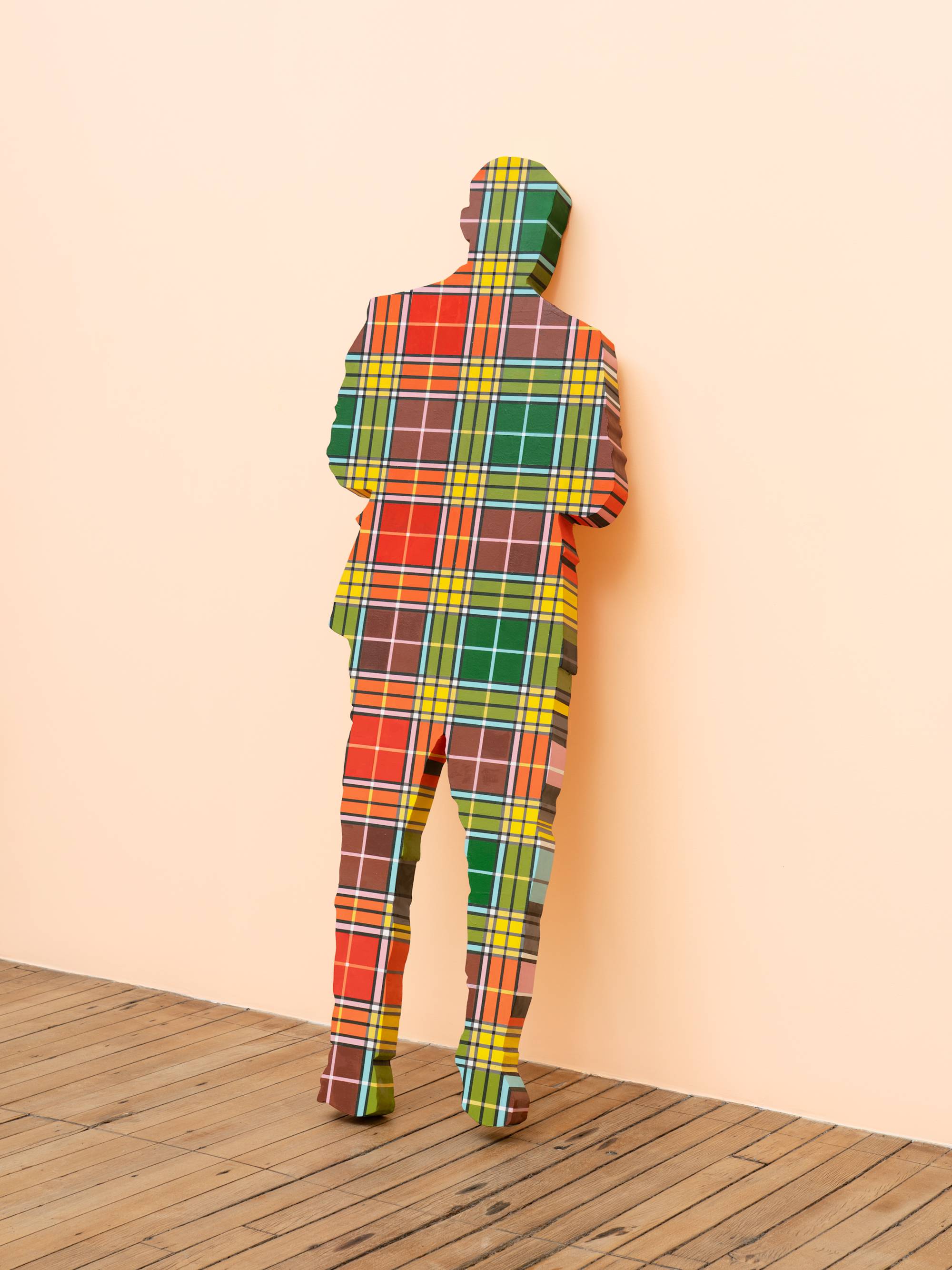 A life-sized human figure painted in a rainbow plaid pattern leans against a peach wall in an art gallery.