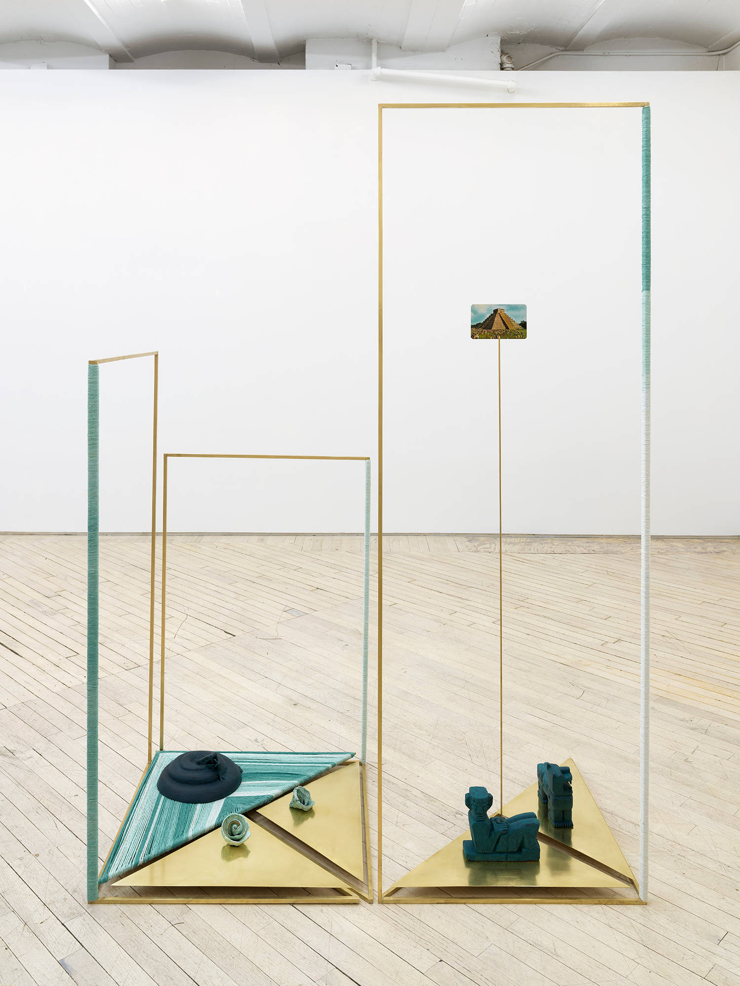 Image of a sculpture by Claudia Peña Salinas composed of vertical brass rods creating rectangular shapes in space with parts of the rod wrapped in dyed blue fabric. Sitting on the base of these rectangles are small sculptures of Mayan symbols, and a postcard stands on a rod of the temple Chichen Itza.