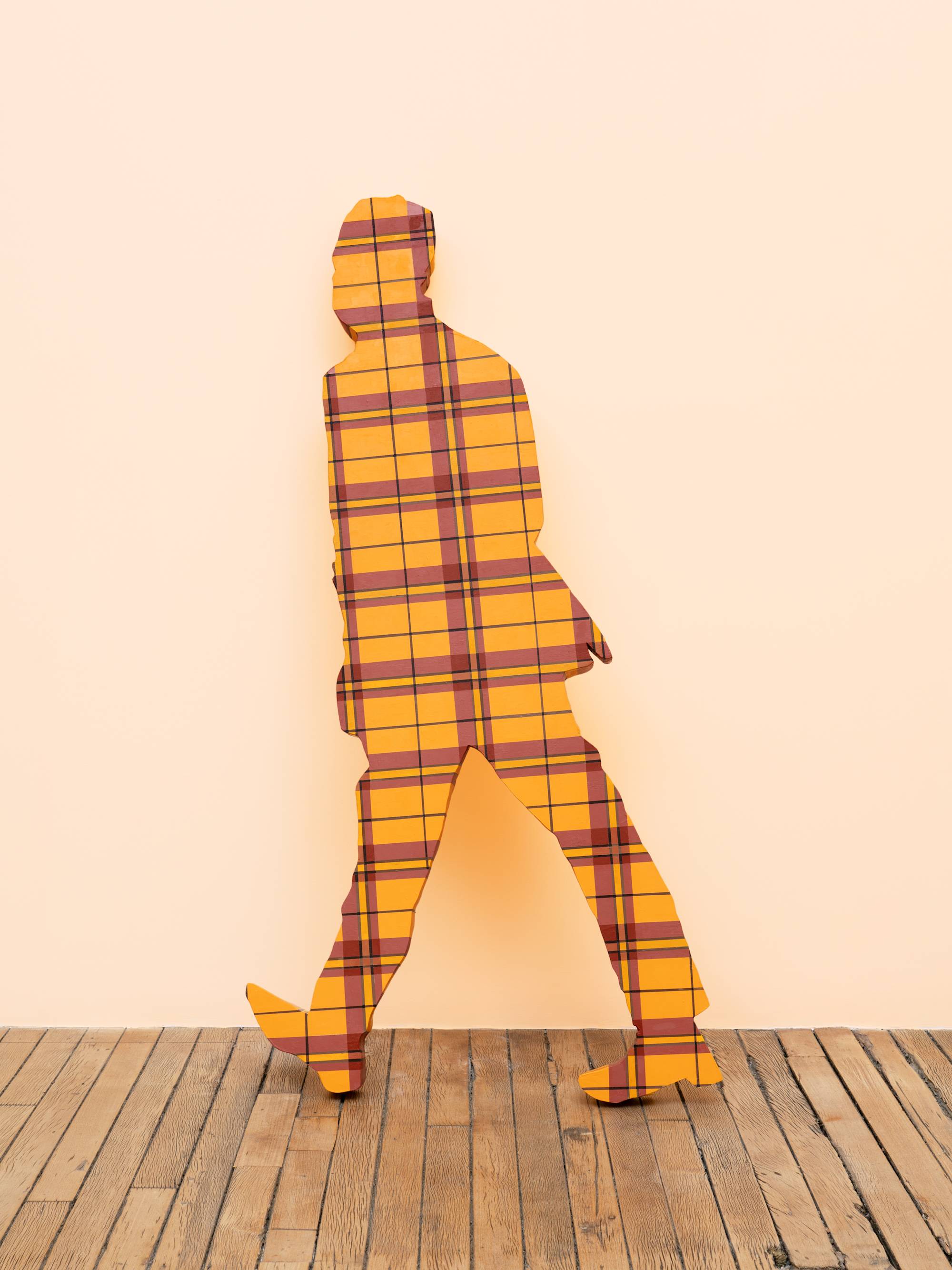 A life-sized human figure painted in a red and yellow plaid pattern leans against a peach wall in an art gallery.