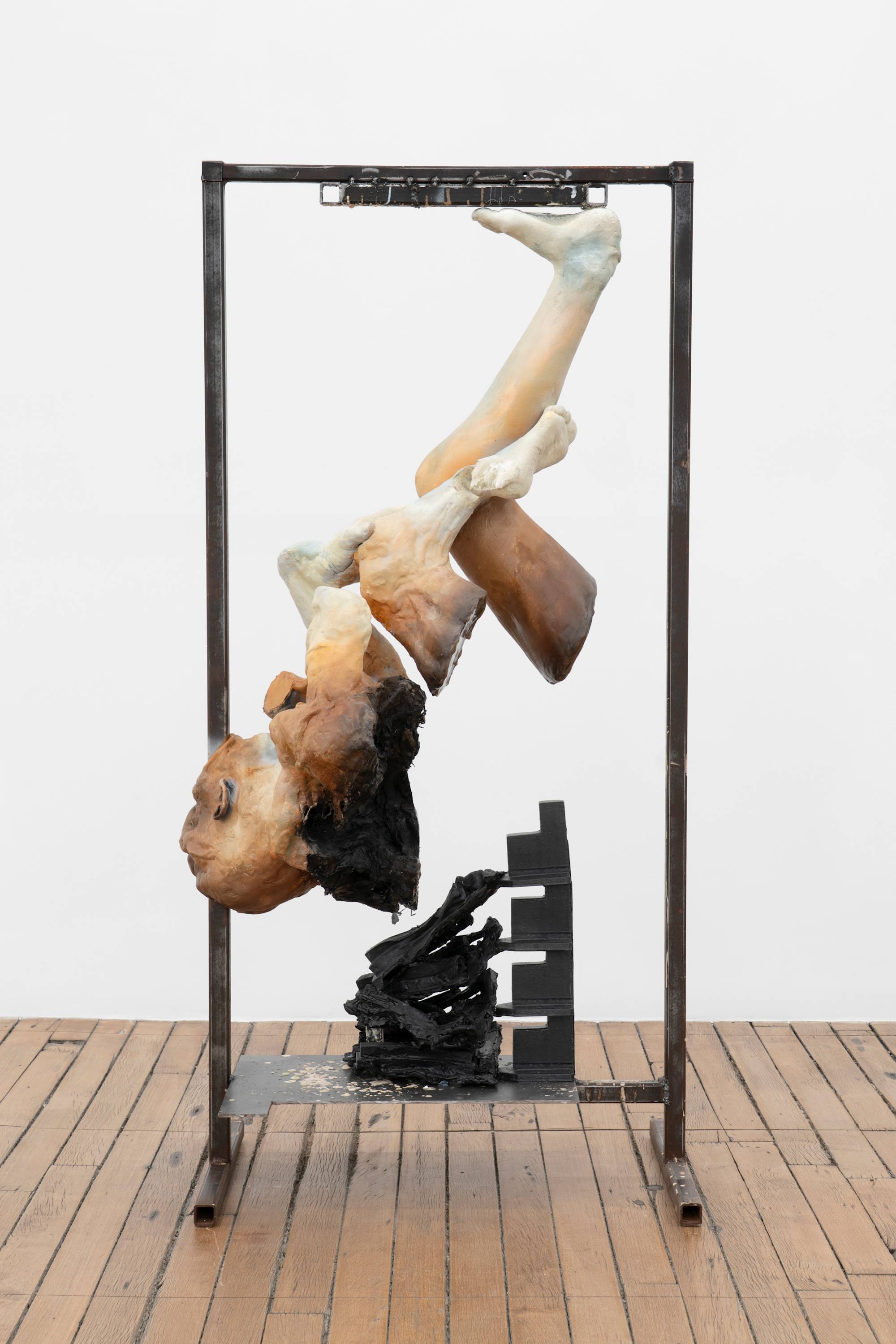 A freestanding sculpture featuring a fragmented figure, inverted and suspended from a metal frame, stands on the wooden floor of an art gallery.