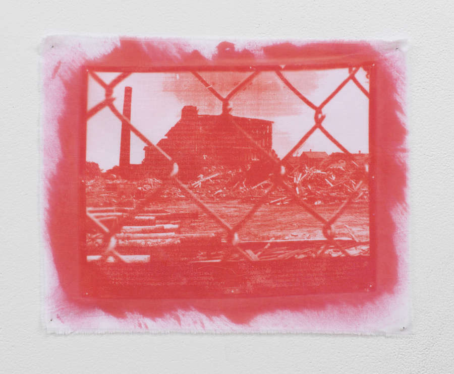 Small photograph of a building behind a chain link fence printed on a cloth in red ink, pinned directly to the wall. 