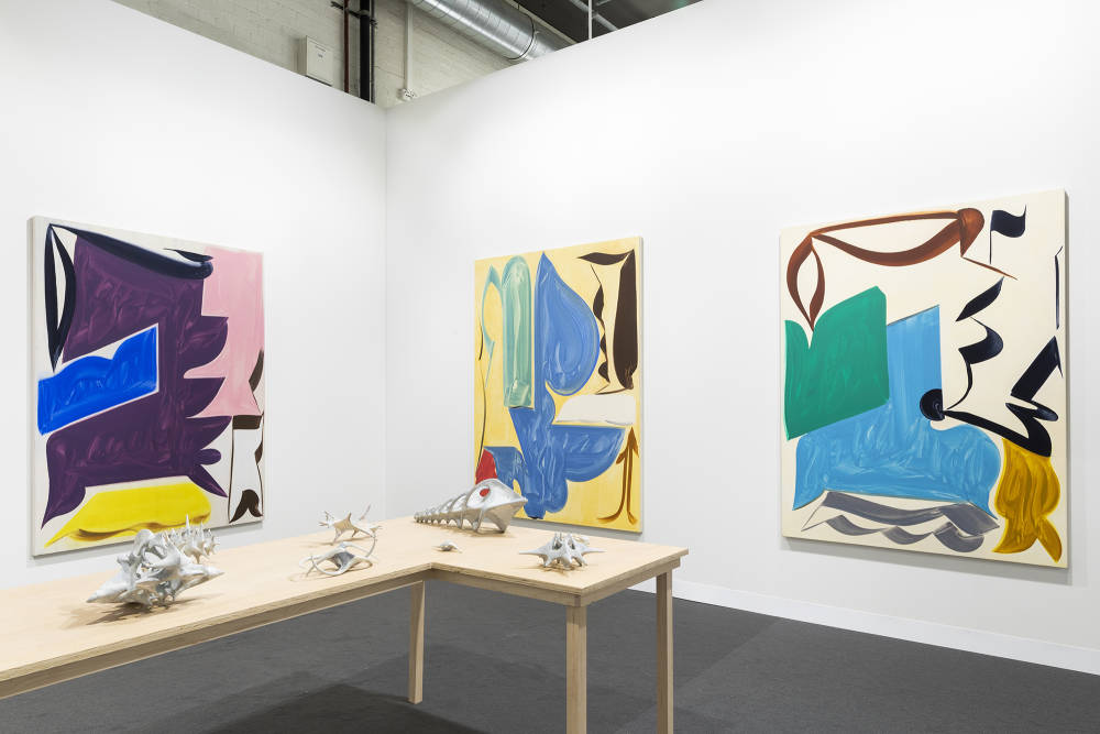 In a gallery space, three large abstract paintings are spaced apart. The painting depict bold abstract geometric shapes in various hues of color. The dominant colors are hues of purple, yellow, blue, pink, brown, and black. The paint is thin and gestural. In the center of the gallery space is a freestanding brownish table with various miniature gray sculptures placed evenly apart. The sculptures are abstract and resemble organic material or bone formations. 