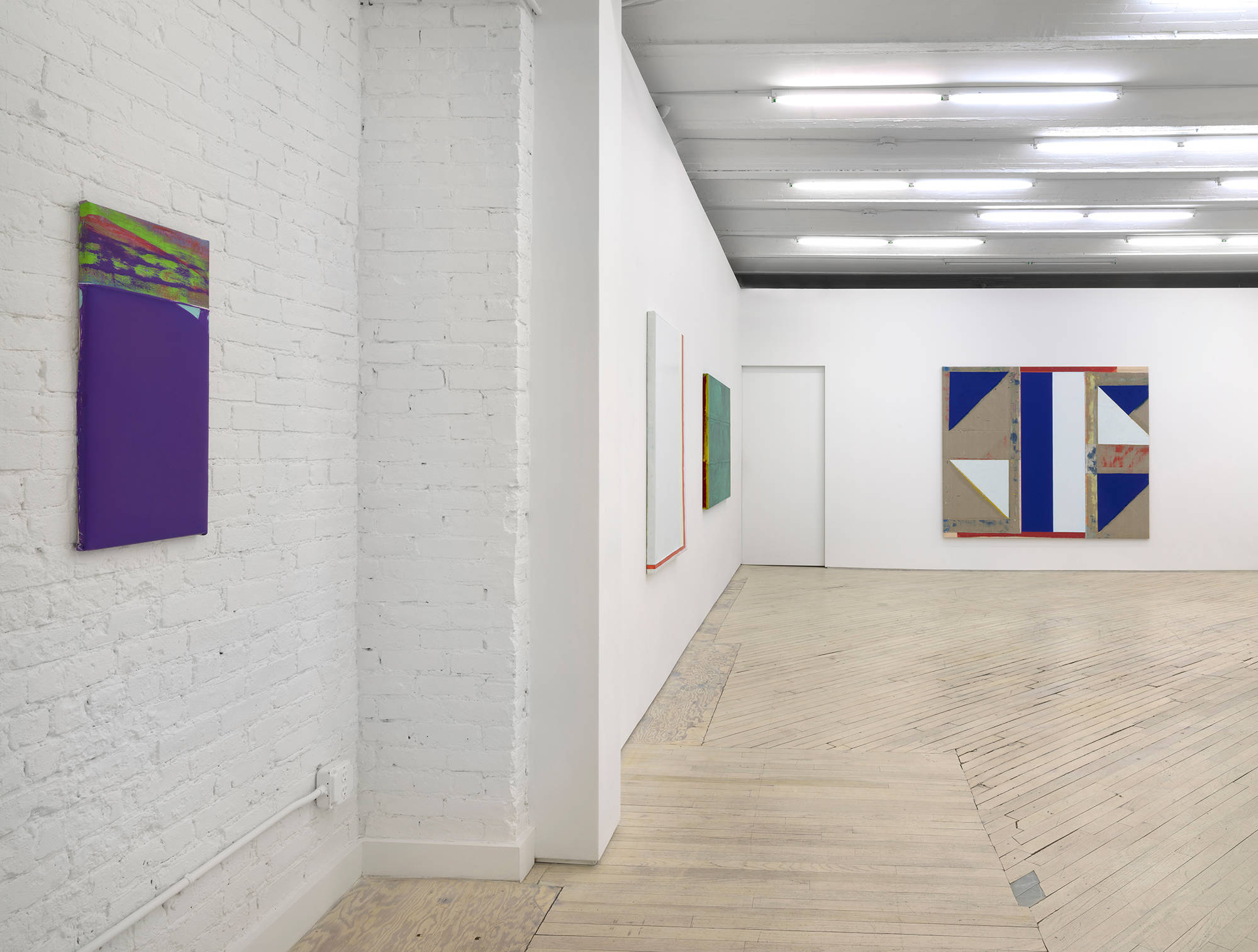 Along a gallery wall, a small purple painting installed next to a larger monochromatic white painting next to a green painting. On the back wall is a larger abstract painting consisting of blue, white, red, and brown areas.