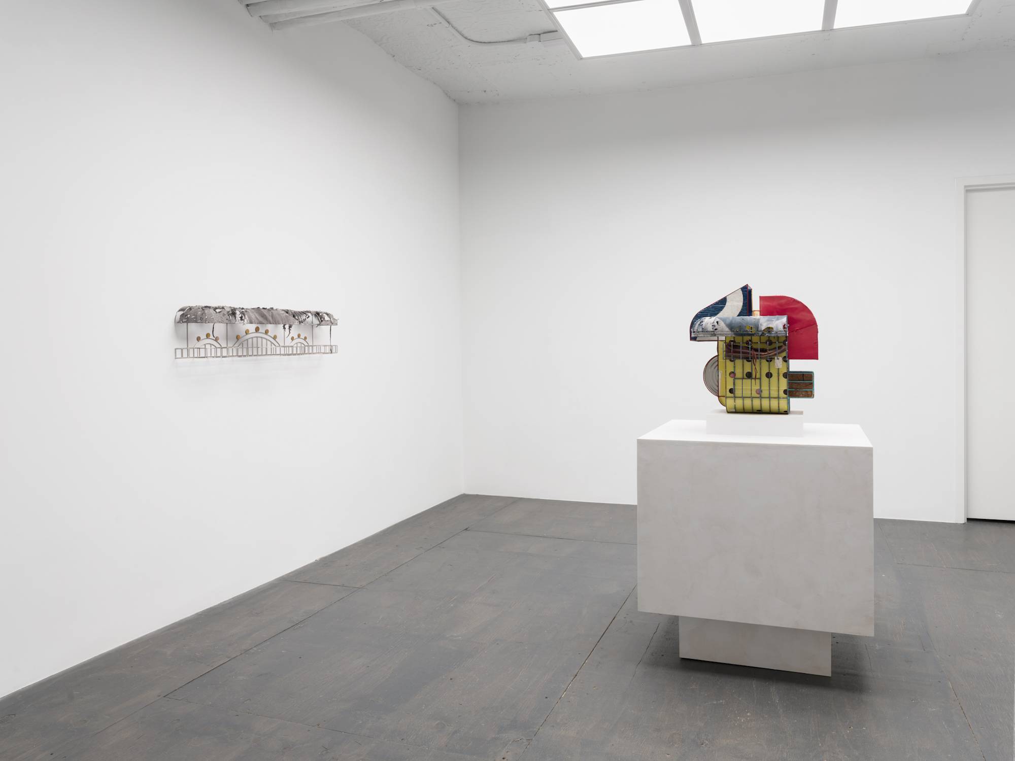Angle view of a white walled gallery with grey floor and white column lit by an LED light fixture that resembles a skylight. Two sculptures are visible in the gallery. One small, silver wall mounted sculpture on the wall to the left and one standing on a pedestal in the center of the room.