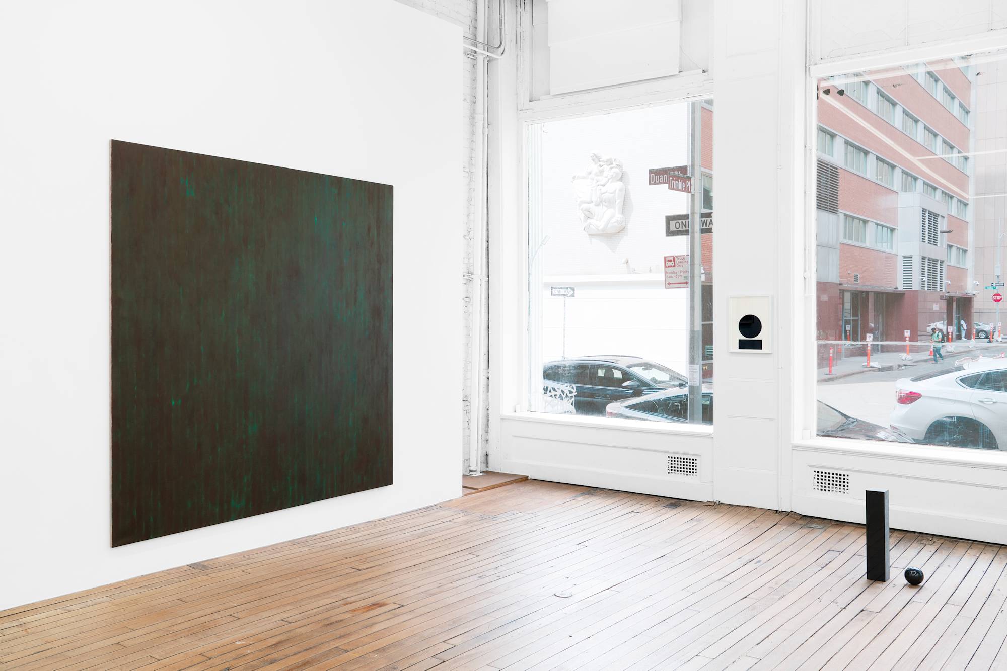 In the corner of an art gallery, on the left wall, a large, abstract painting with brown and green tones hangs. A framed drawing of a cropped "i" hangs on a narrow section of wall between two large windows which look out upon the street. On the floor, to the right of frame, sits a sculpture of an "i" that's tittle has fallen out of suspension and to the floor.