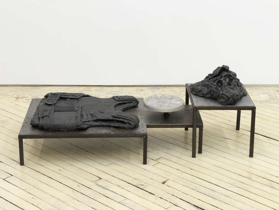 In a gallery space, an iron sculpture depicting abstracted forms on top of a plinth.