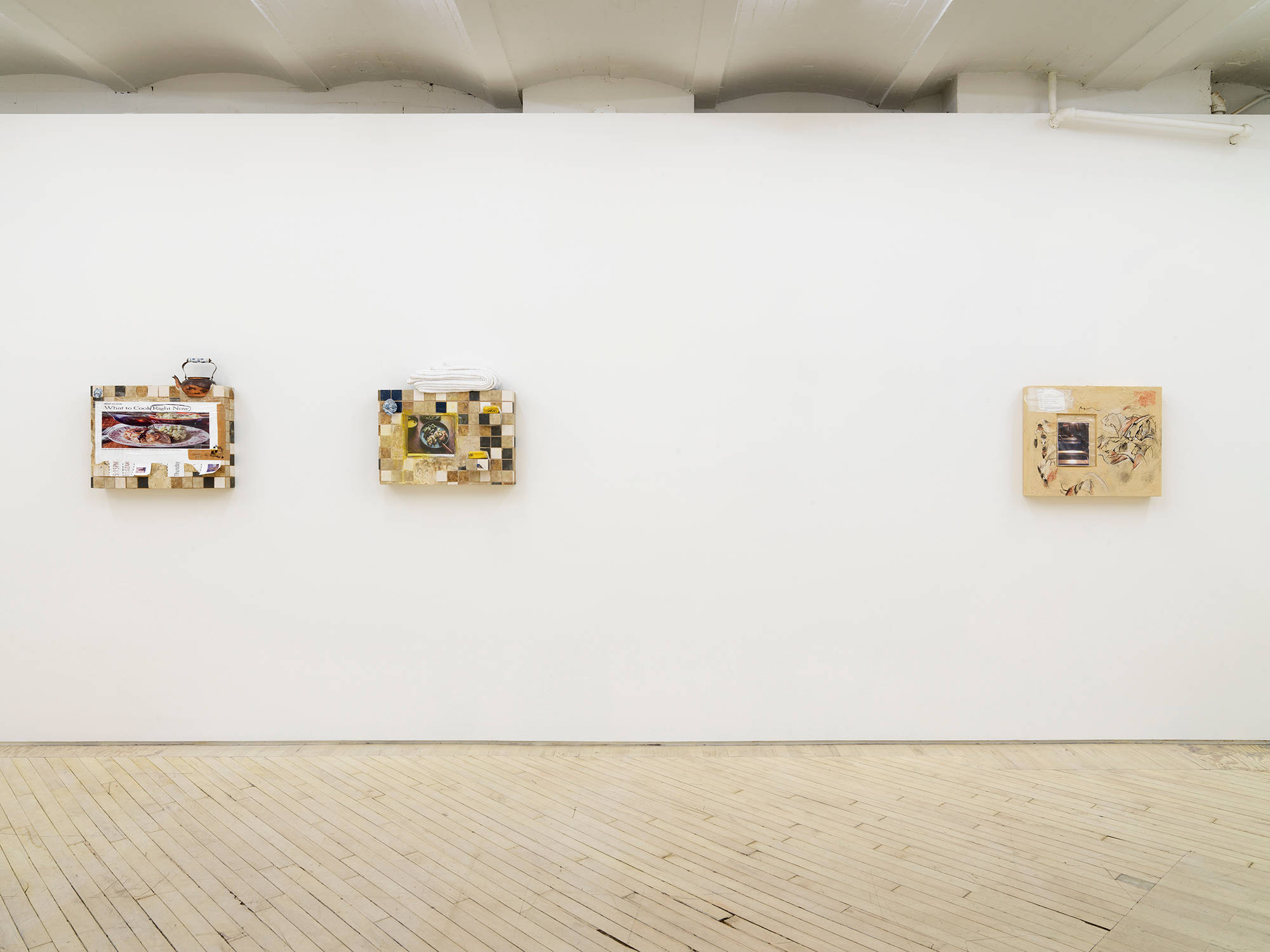 On a long white gallery wall are three wall sculptures. Two of the sculptures are spaced closed together on the left. They are rectangular and resemble arbitrary patterns of kitchen titles with images adhered to them. The piece on the right is also rectangular and resembles a handmade cave drawing with another printed photograph adhered to it.