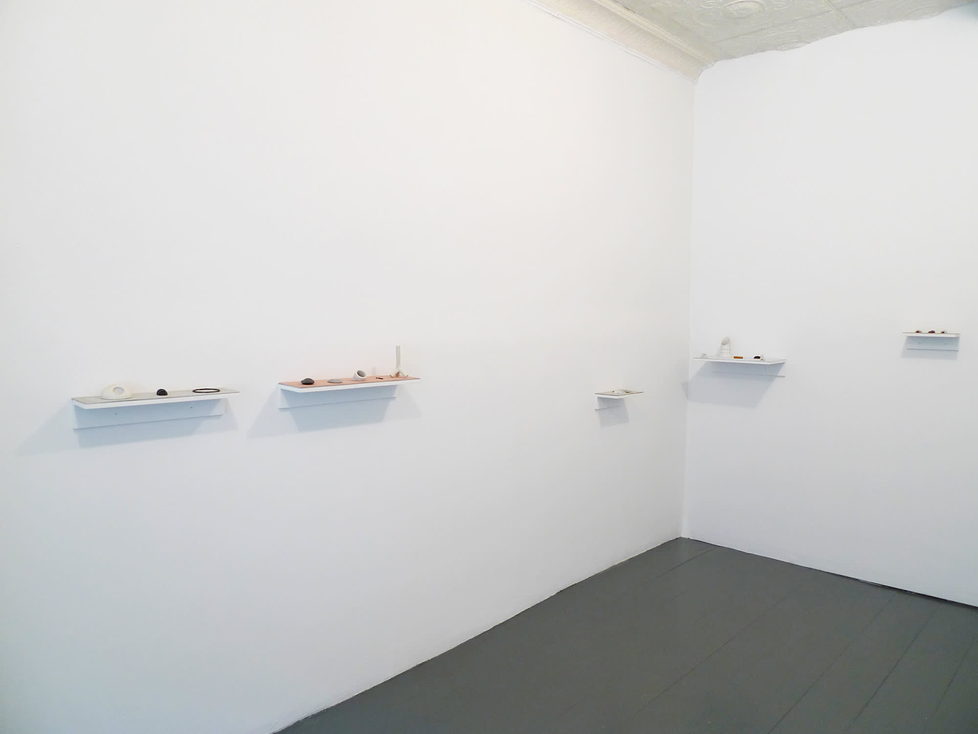In a gallery space, numerous small white shelves are installed throughout the room with multiple small objects resting on top of them.