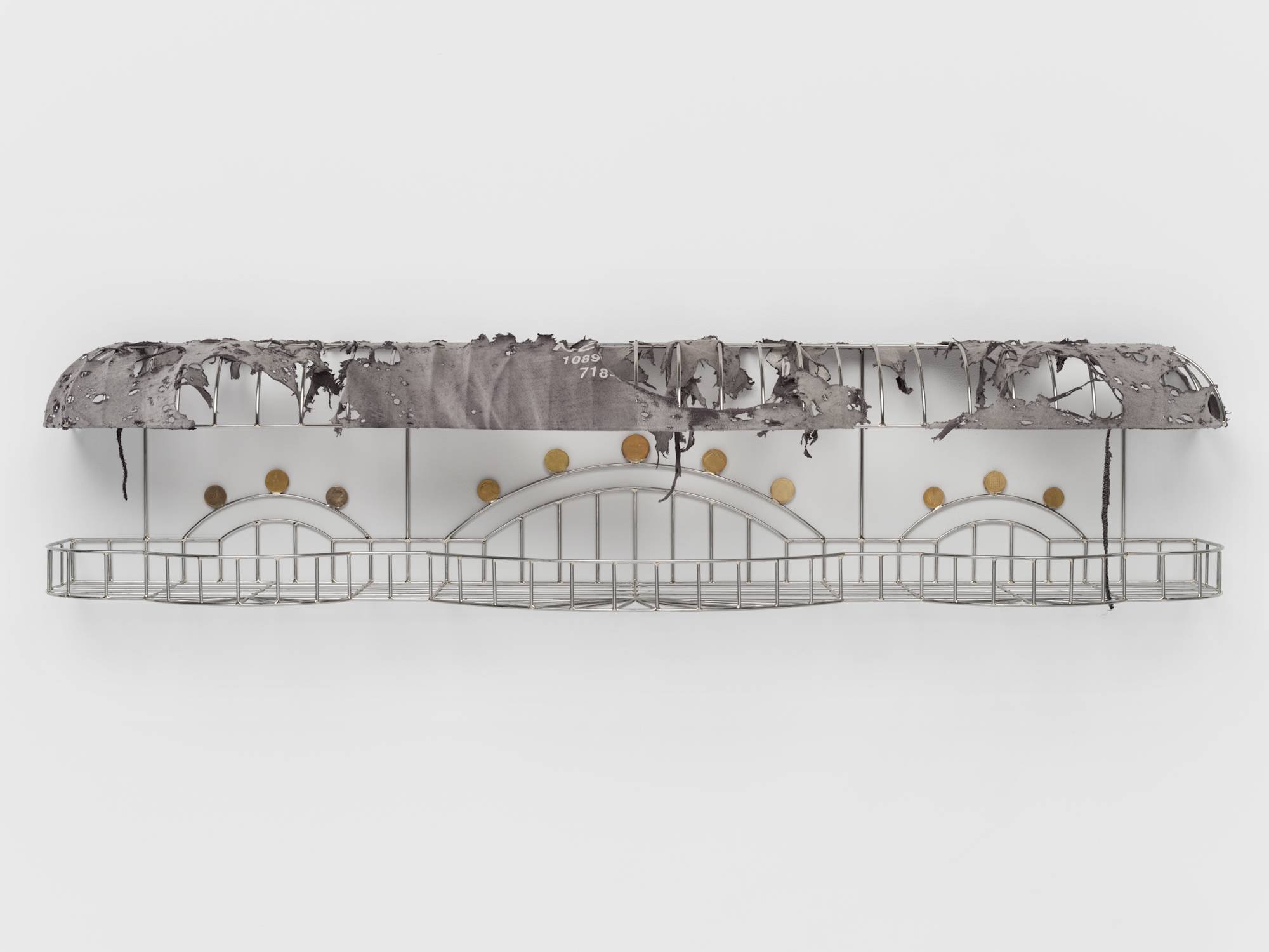 A cage-like chrome sculpture, wrapped with a tattered grey t-shirt to resemble an awning, and embellished with soldered on gold-colored coins hangs on a white wall.