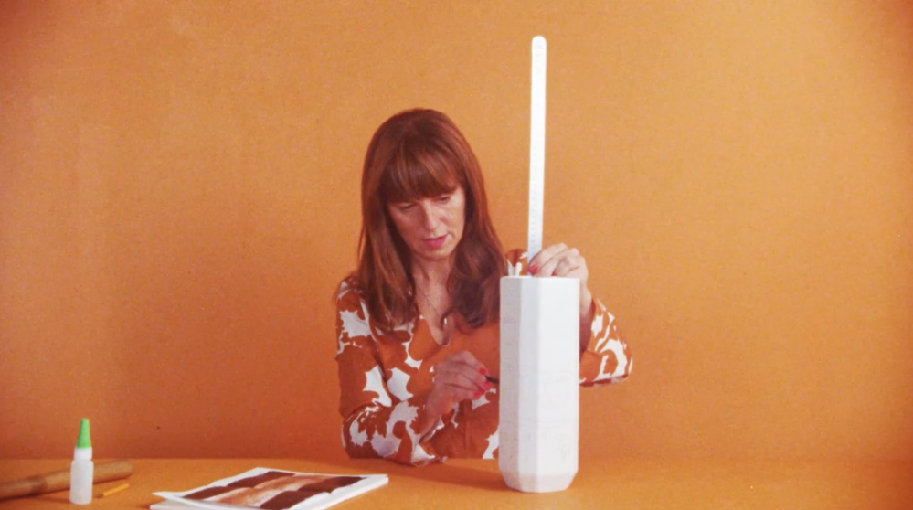 Still image from the film The Uruk Vase showing a seated woman in an orange shirt against an orange colored background measuring a vase with a measuring stick