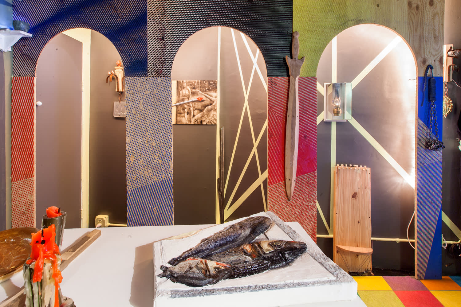 A camera image of a colorful room with three archways that show a shallow lit space with images on the wall, and simple painted mural of lines. A large sword hangs between the right two archways. A wooden sculpture sits in the third archway. In the front of the image is a distorted sculpture, implying a specific perspective, of cooked fish on a platter next to two candles.