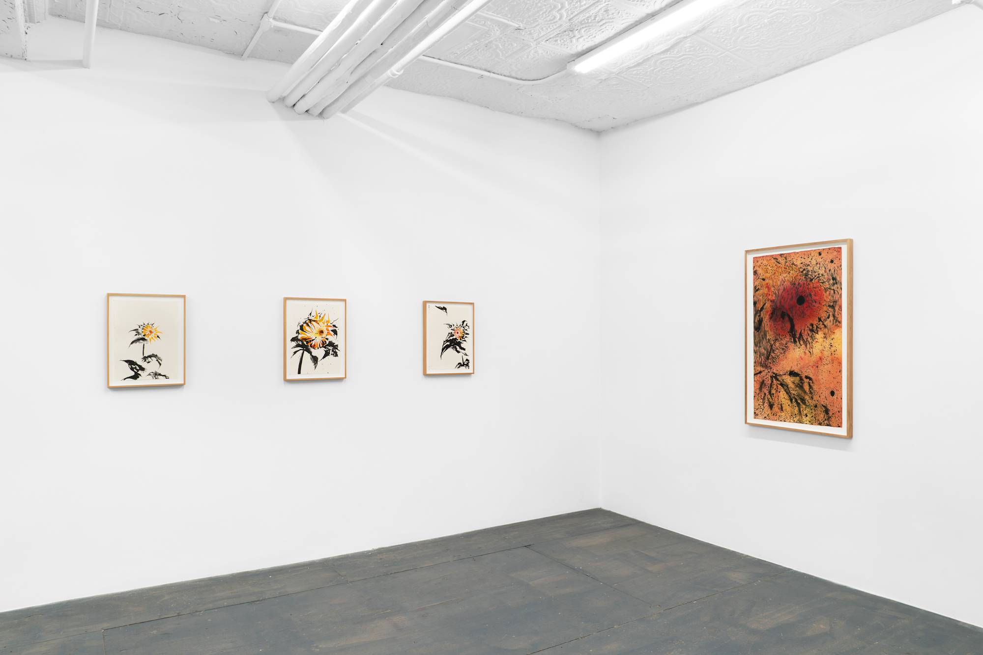 An interior shot of two white walls inside a gallery space: on the left wall, three small yellow-and-black ink paintings of sunflowers are mounted in a horizontal row; on the right wall, a larger red, orange, and black in painting of a sunflower is mounted.