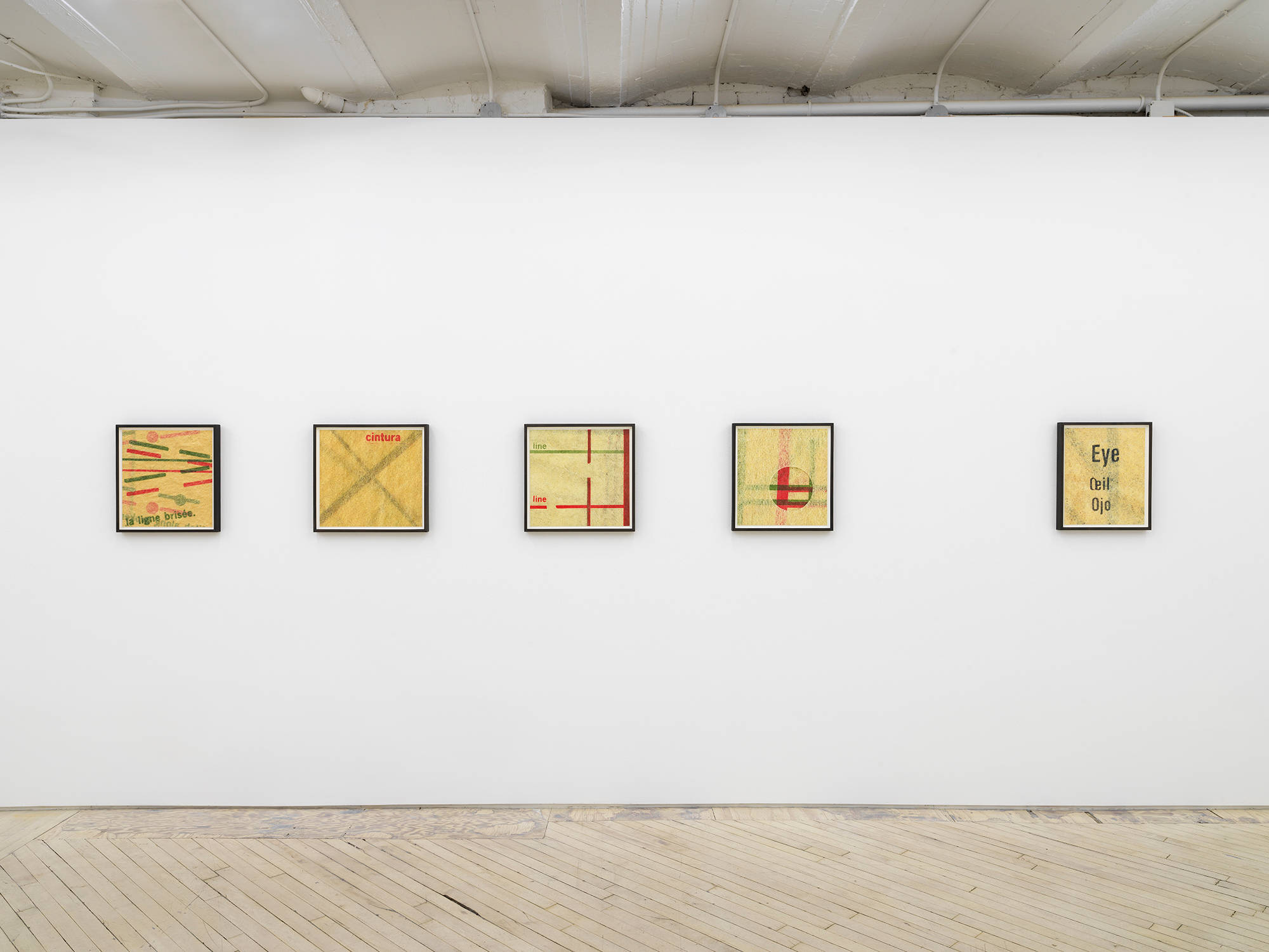 In a gallery a series of photographs in a black frames hang in a line on a wall. The photograph are depicting sewing patterns with aged and yellowed paper.