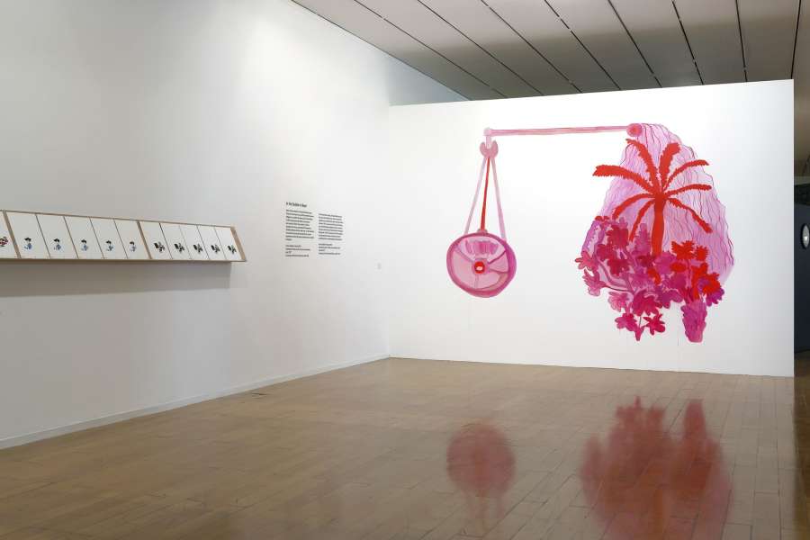 In a large gallery space a large abstract wall drawing depicting a pink form. To the left a series of drawings hung in a display case.
