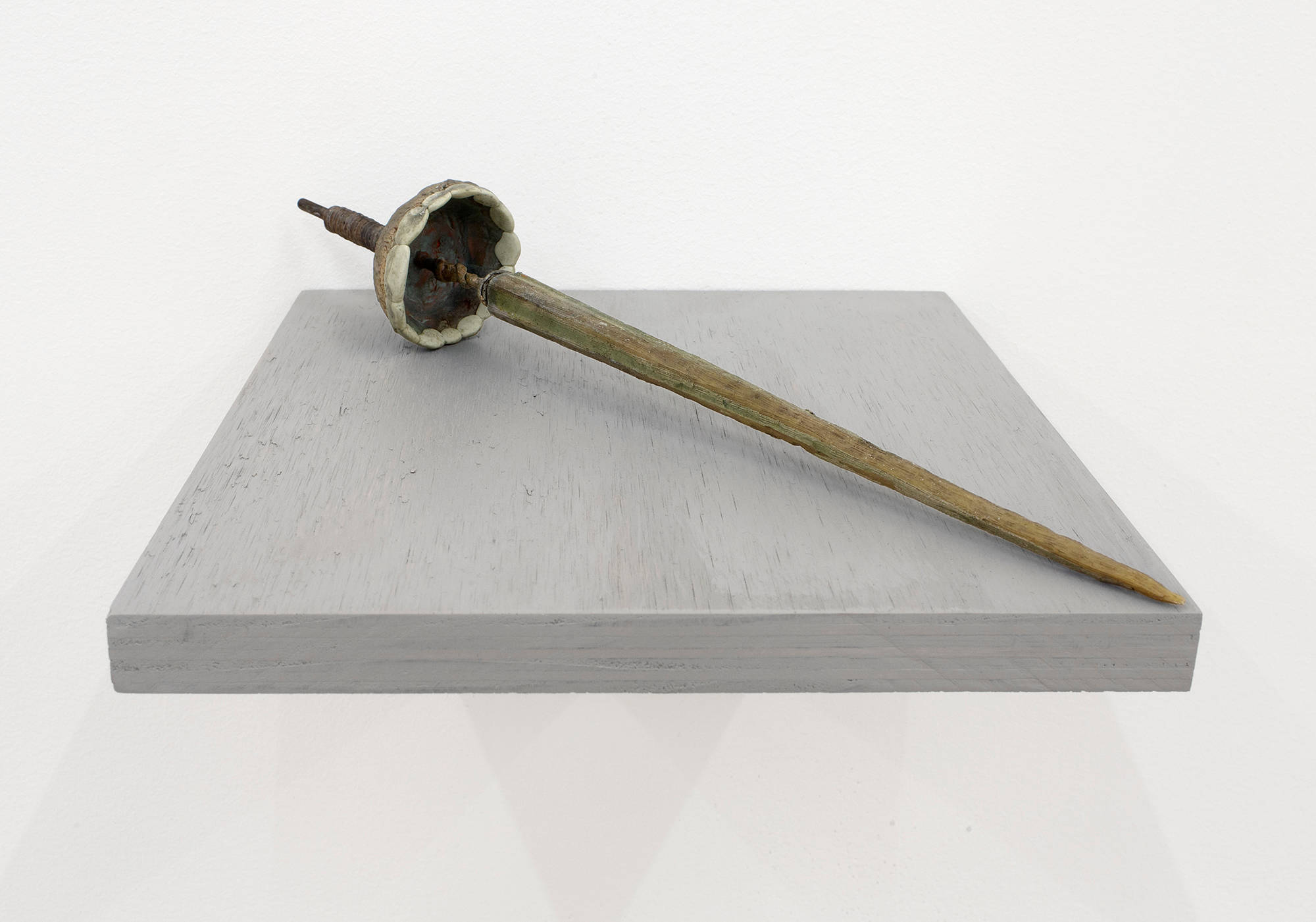 A close-up of a handmade sculpture resembling an ancient pointy tool with a long handle. The sculpture is resting on a gray shelf installed directly to the wall.