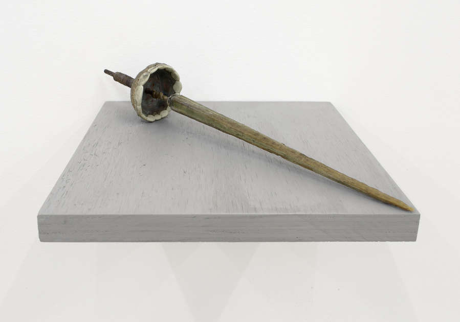 A close-up of a handmade sculpture resembling an ancient pointy tool with a long handle. The sculpture is resting on a gray shelf installed directly to the wall. 