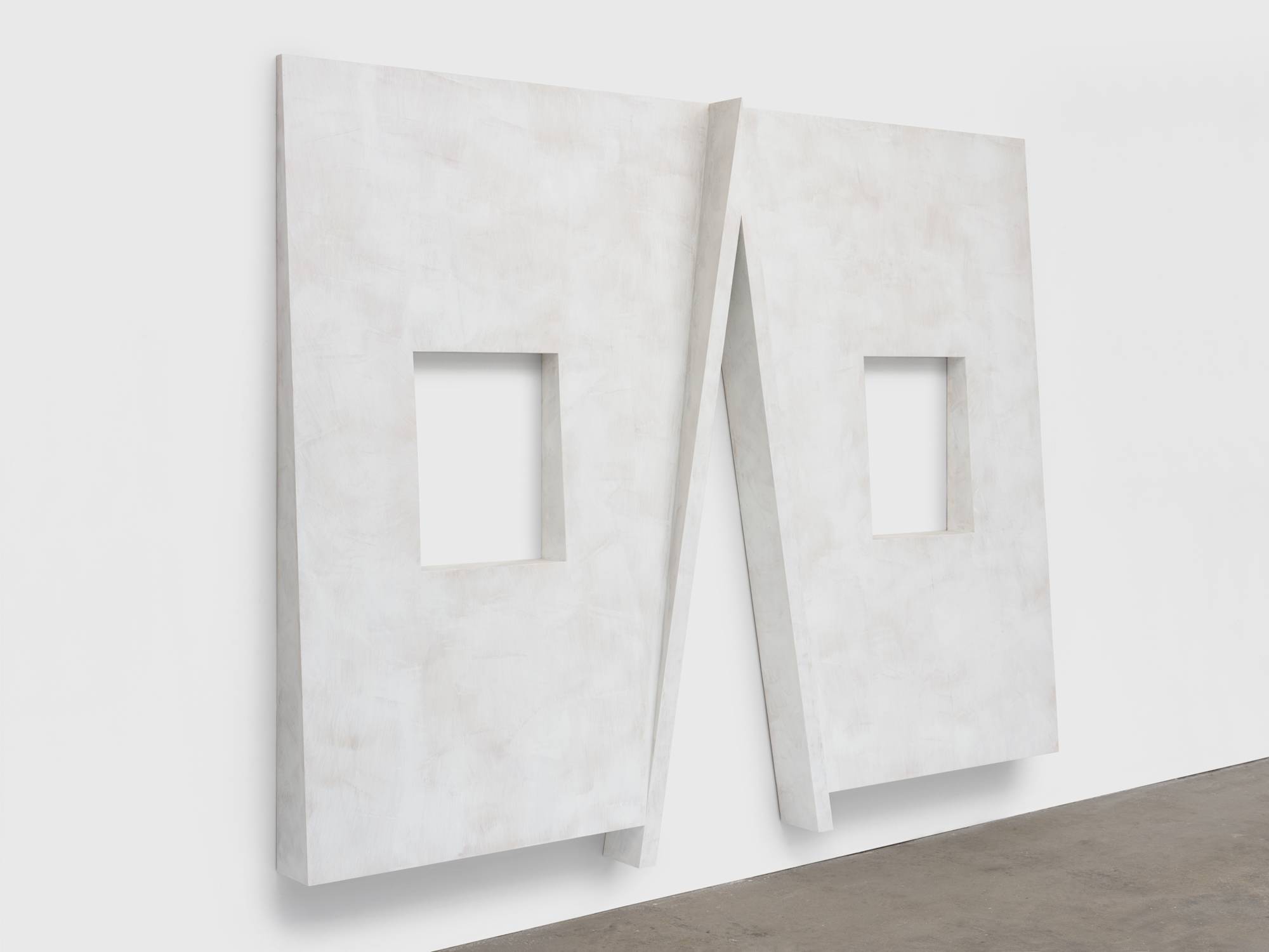 Angle view of a large wall sculpture designed with a gently receding slope from bottom to top with two rectangular window cutouts on either side hanging on a white wall above a grey floor. The center features a cutout resembling a cross gable roof.