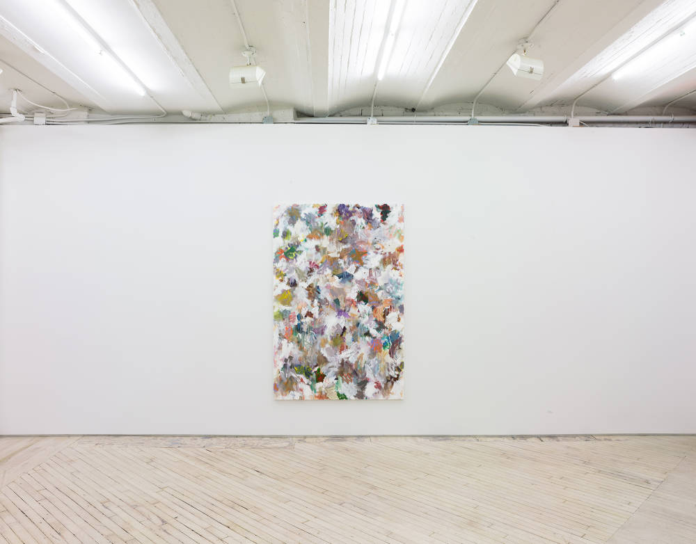 In a gallery space, a large abstract paintings is hung in the center of a large white wall. The painting contains numerous swatches of various colors painted on top of one another. The paint is thin and in some areas dripping.
